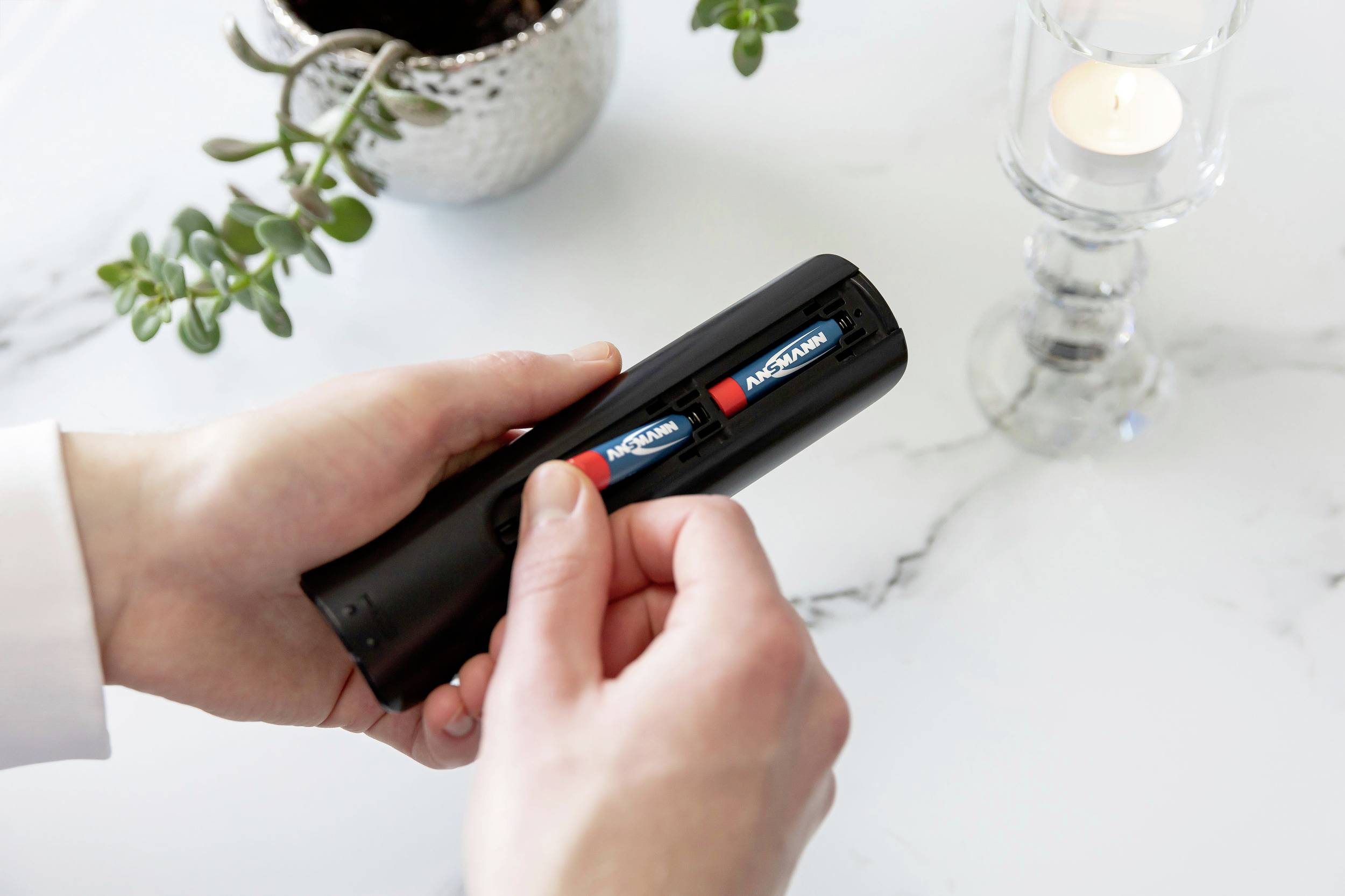 Person legt zwei Batterien in eine Fernbedienung ein. Auf einem Tisch steht eine brennende Kerze und eine Pflanze als Dekoration.