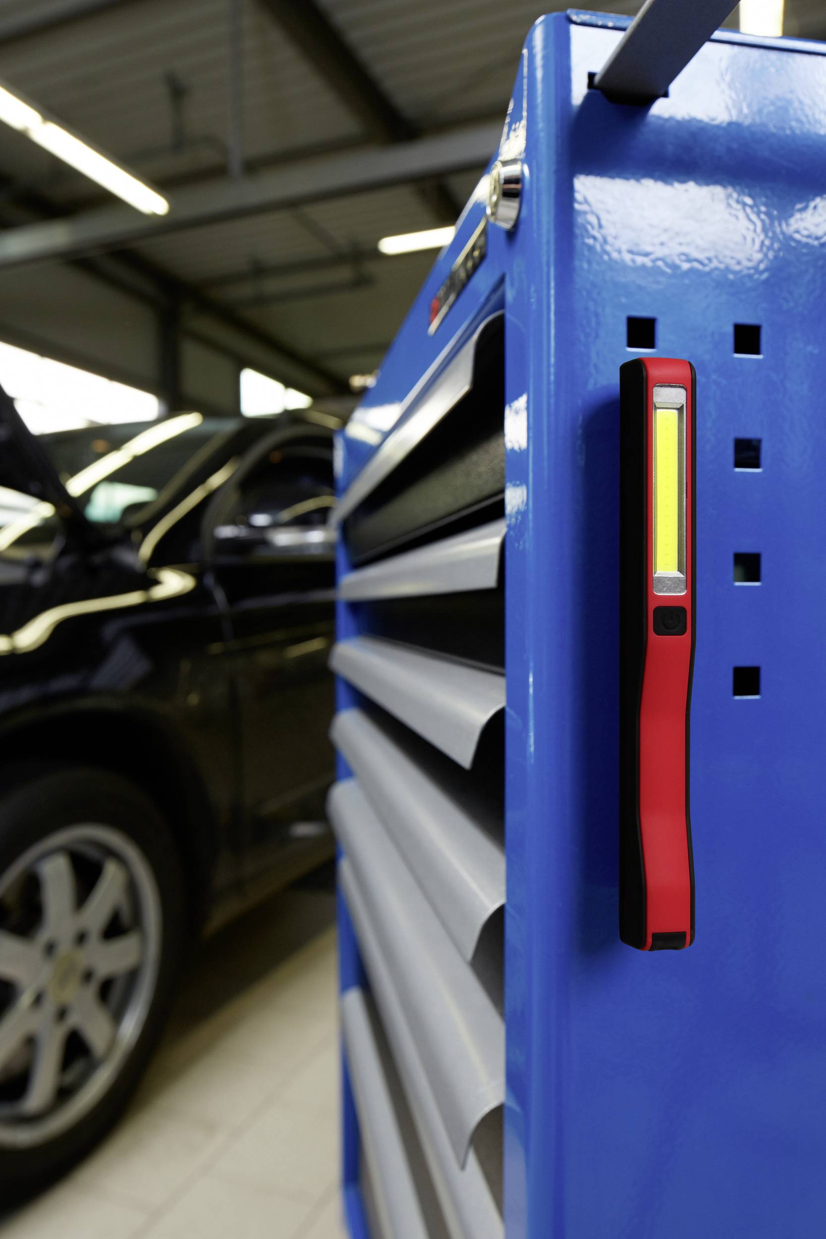 Ein blauer Werkzeugschrank mit einer magnetischen roten Taschenlampe an der Seite. Im Hintergrund steht ein offenes Auto in einer Werkstatt.