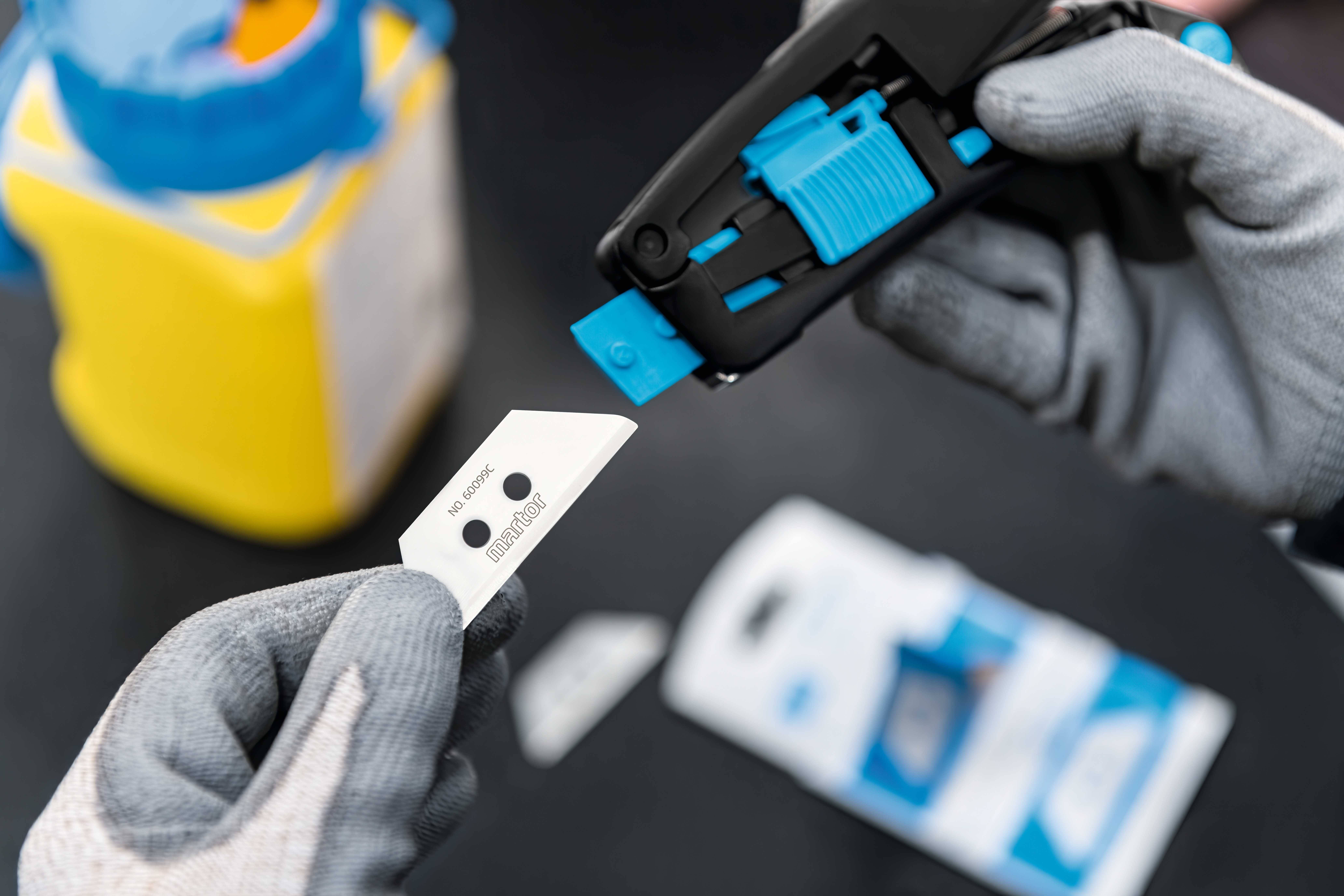 Hand zieht Klinge aus einem schwarzen Klingenabbrecher, im Hintergrund verschwommenes gelbes Objekt. Thema: sicherer Klingenwechsel.