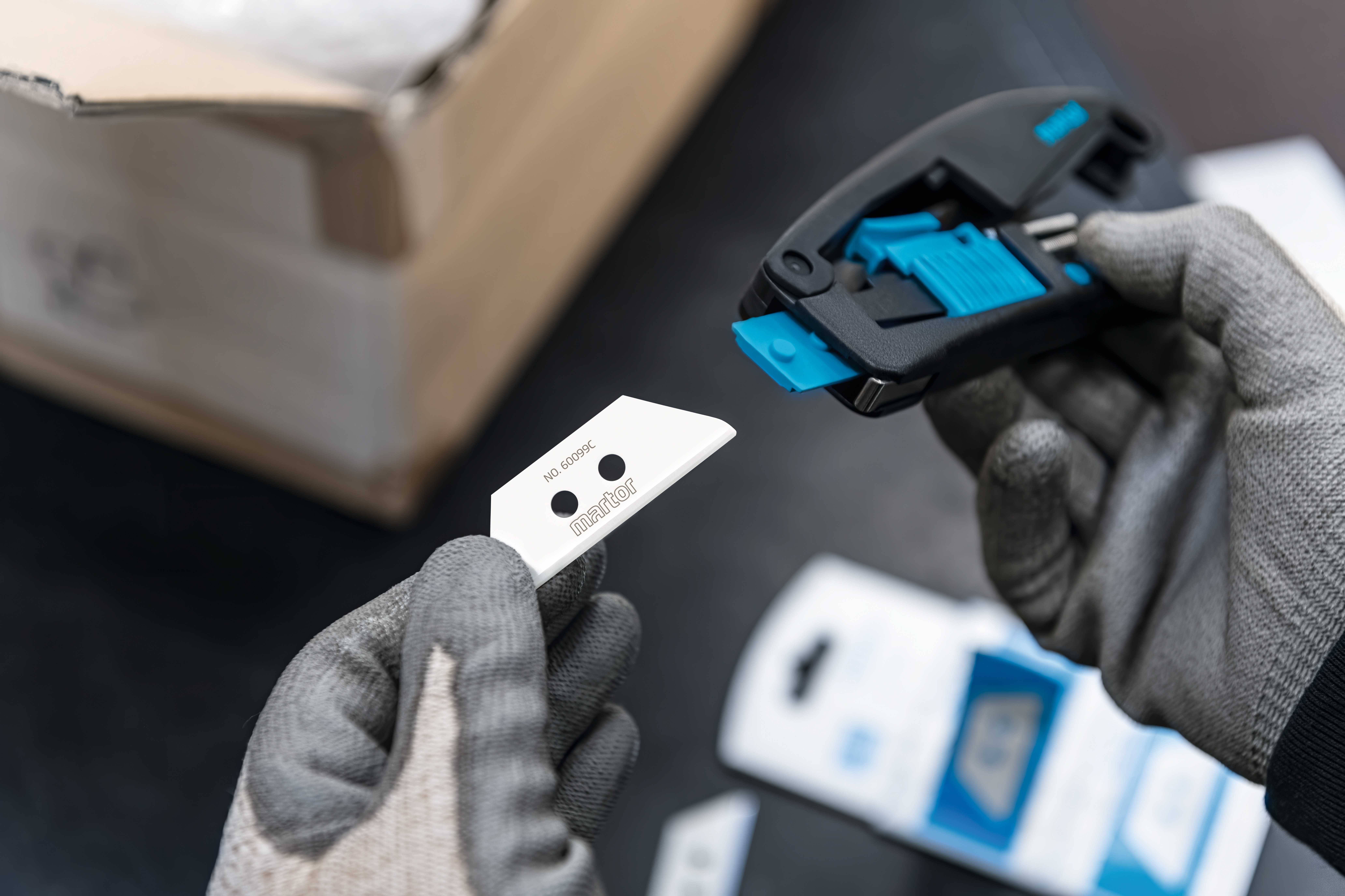 Eine Person mit Handschuhen hält einen Ersatzklingenhalter und eine neue Klinge, bereit zum Einsetzen in den Halter. Im Hintergrund ist ein Karton.