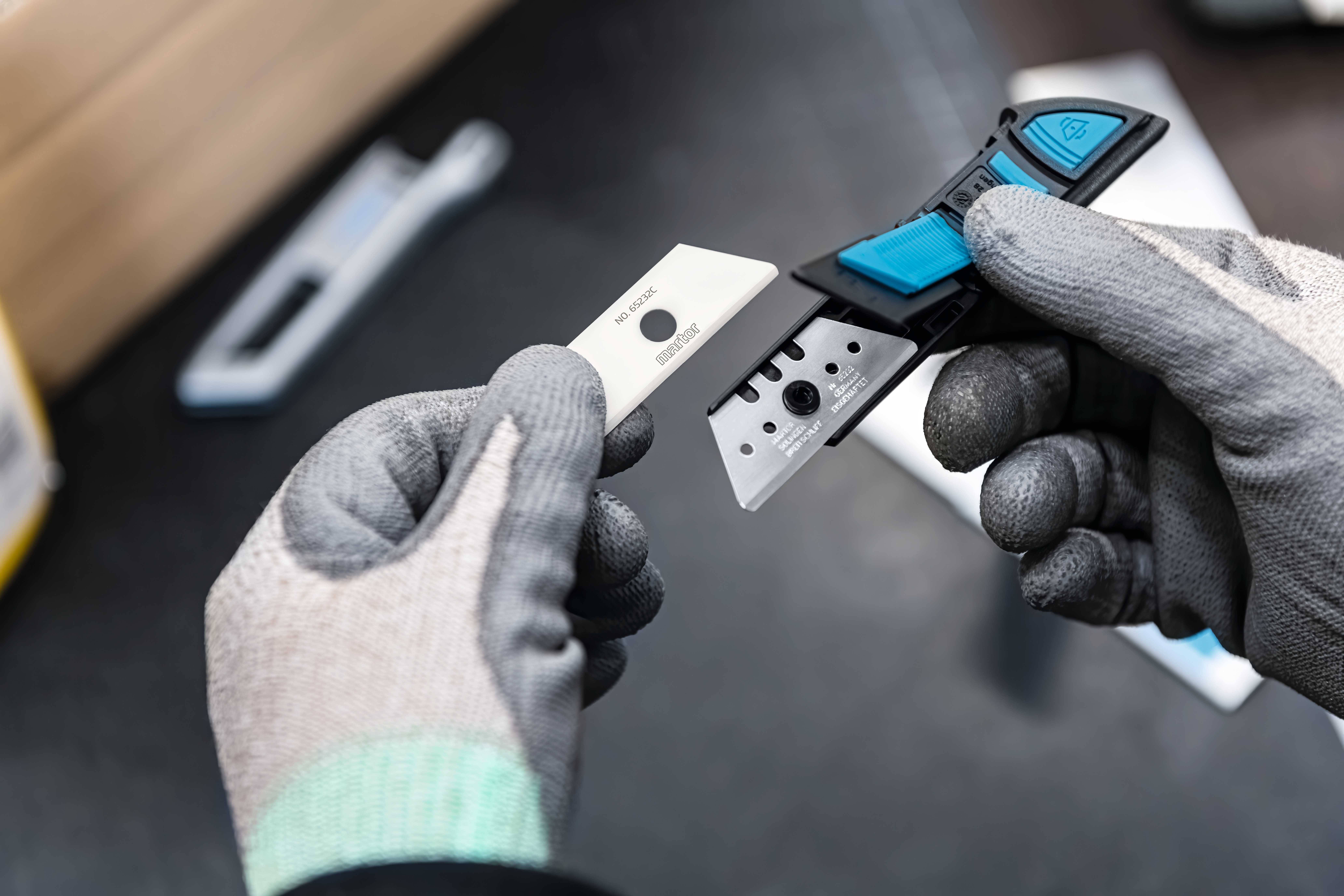 Eine Person mit Handschuhen hält ein Messer mit einem neu eingesetzten Ersatzklingenfach. Im Hintergrund ist unscharf weiteres Werkzeug zu sehen.