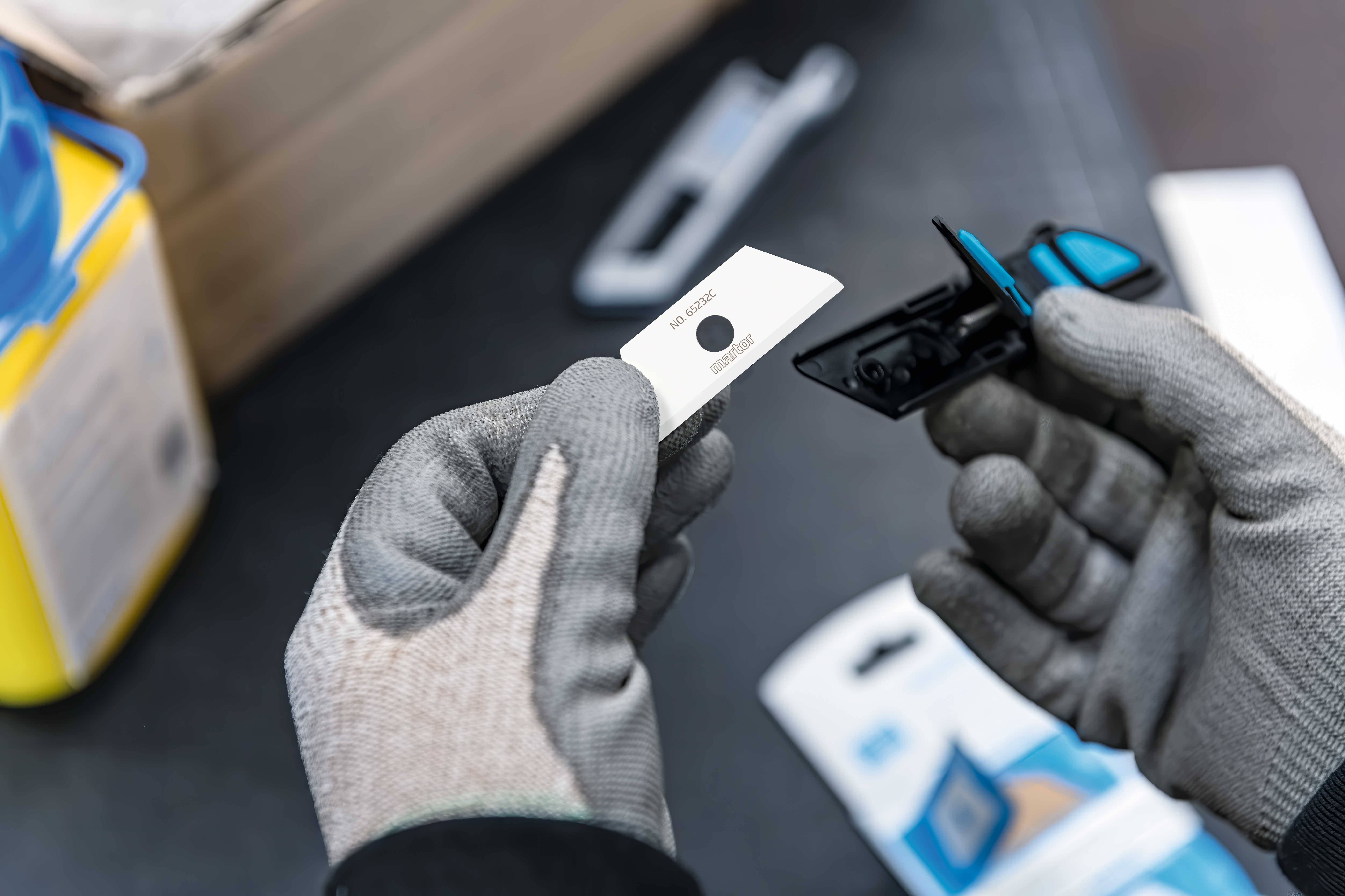 'Hand mit Handschuh hält eine Rasierklinge neben einem Rasierer. Im Hintergrund sind eine Klinge in einer blauen Halterung und Verpackungen erkennbar.'