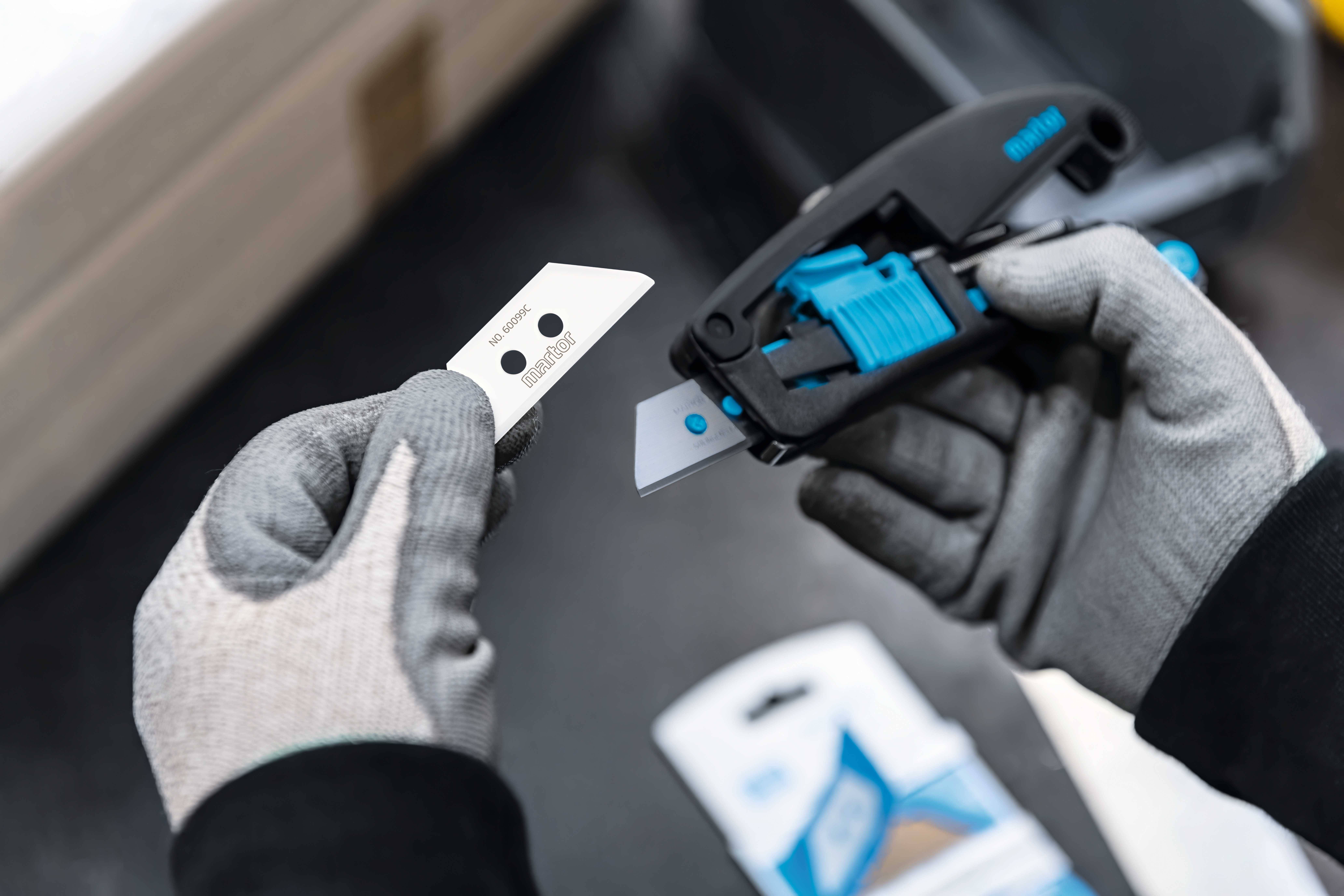 Eine Person mit grauen Handschuhen wechselt das Klingenblatt eines Sicherheitsmessers. Im Hintergrund sind Verpackungen sichtbar.