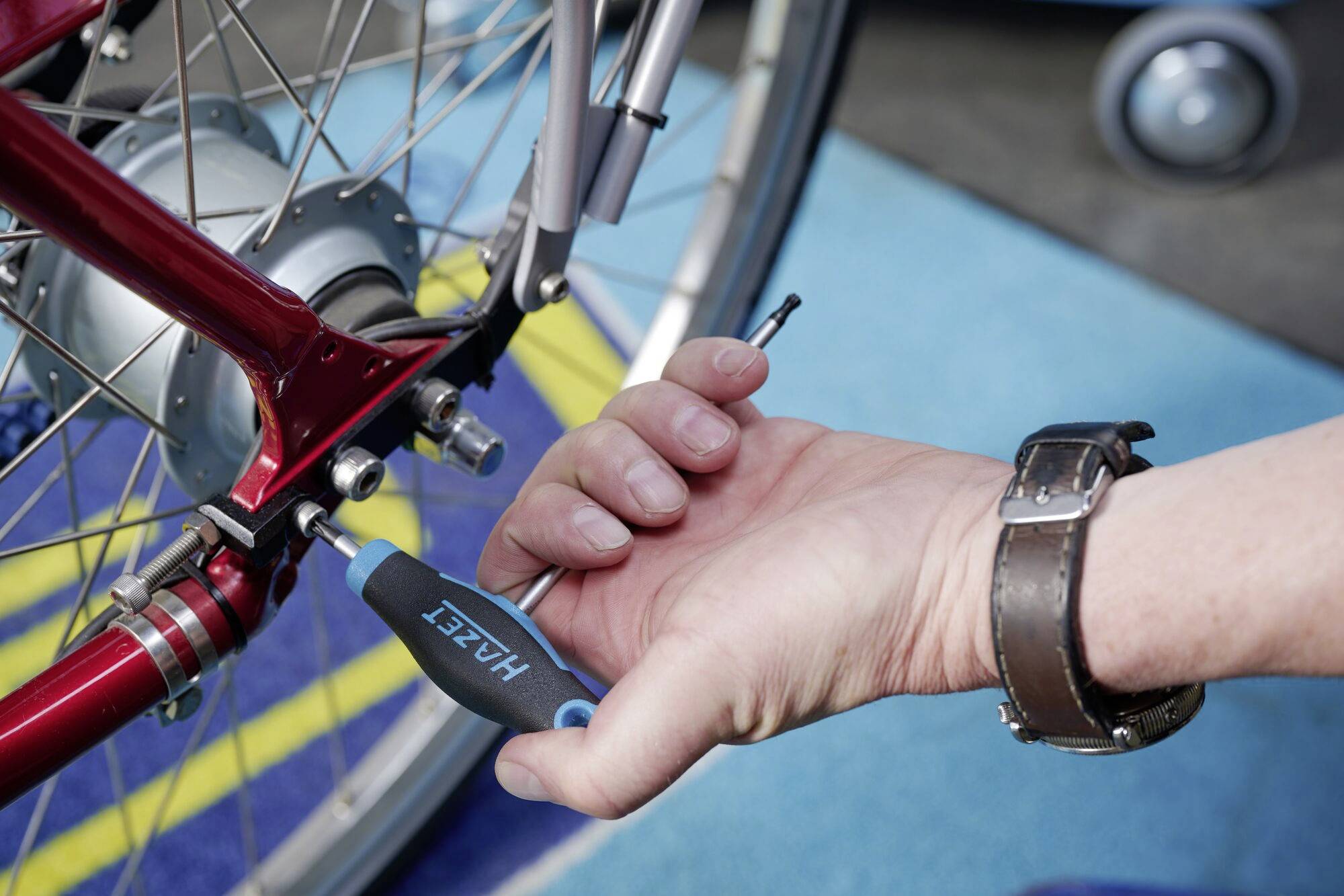 Eine Hand zieht eine Schraube an einem Fahrrad mit einem Schraubendreher fest. Das Fahrrad hat ein rotes Rahmendesign und silberne Speichen.
