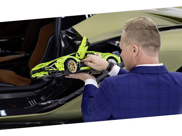 Ein Mann in einem blauen Anzug betrachtet ein Modellauto in Hellgrün vor einem größeren Auto.