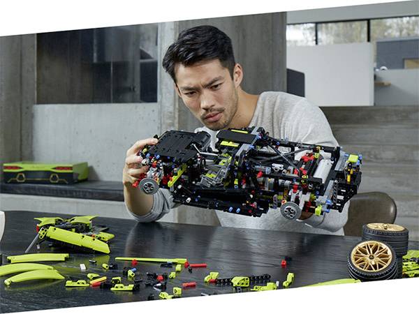 Ein Mann baut ein komplexes Modellauto aus bunten Bauklötzen. Neben ihm liegen Einzelteile und Radkomponenten auf einem Tisch.