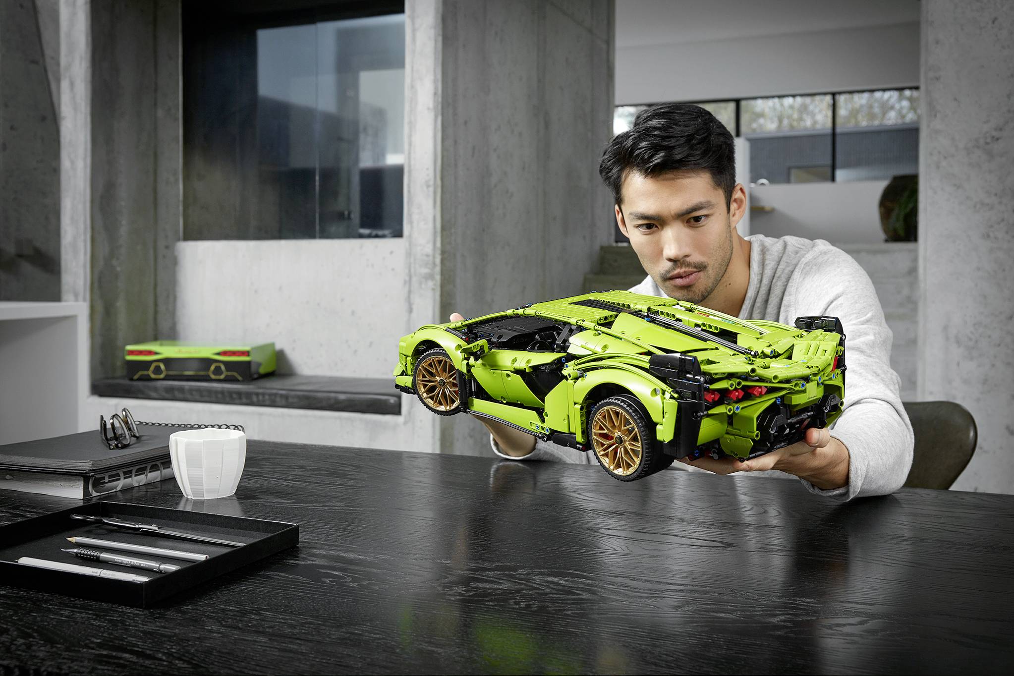 Ein Mann sitzt an einem Tisch und hält ein grünes Modellauto. Im Hintergrund steht ein weiteres Modellauto.