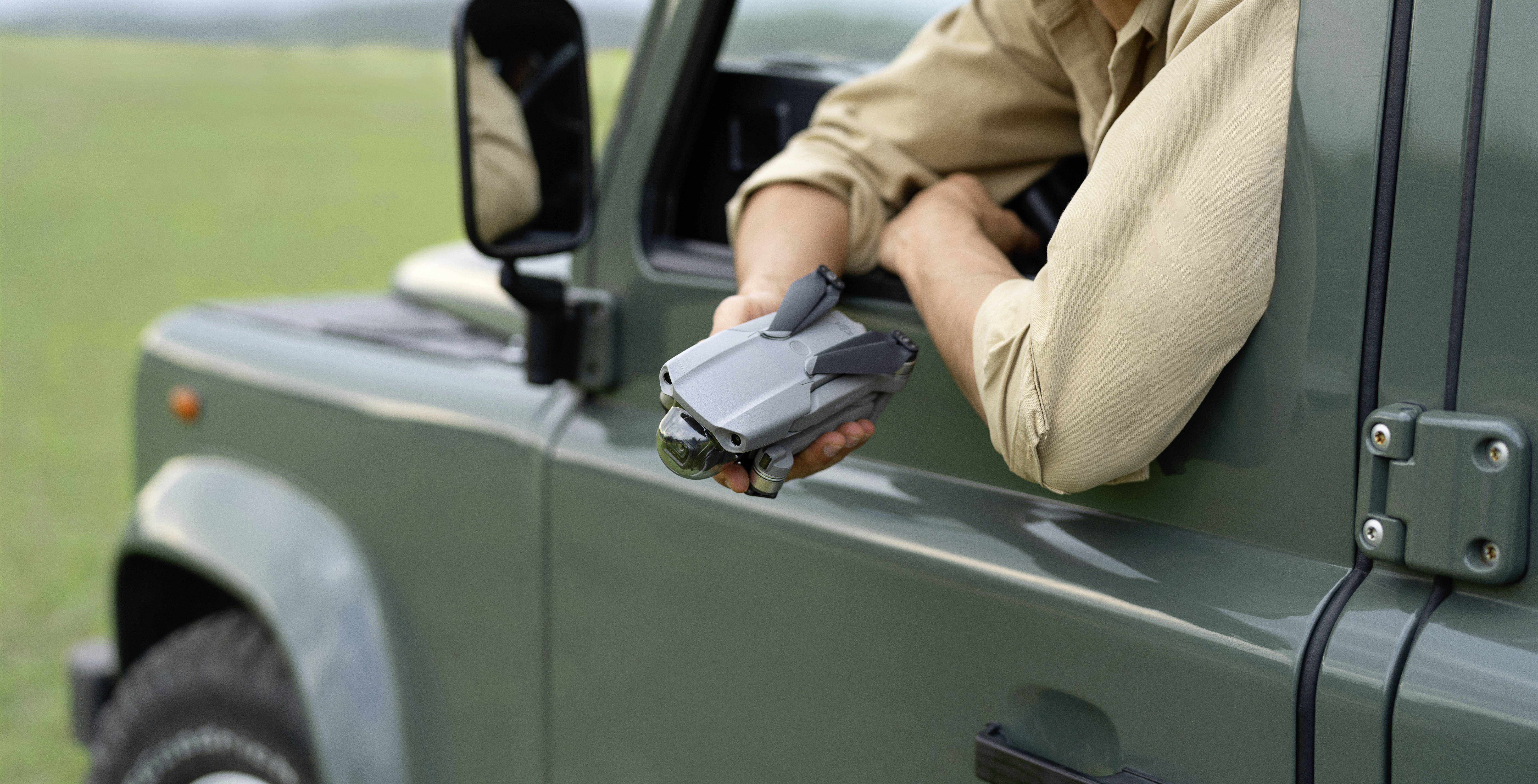 Person hält eine Drohne aus einem Autofenster, bereit zum Starten, vor einer grünen Wiesenlandschaft im Hintergrund.