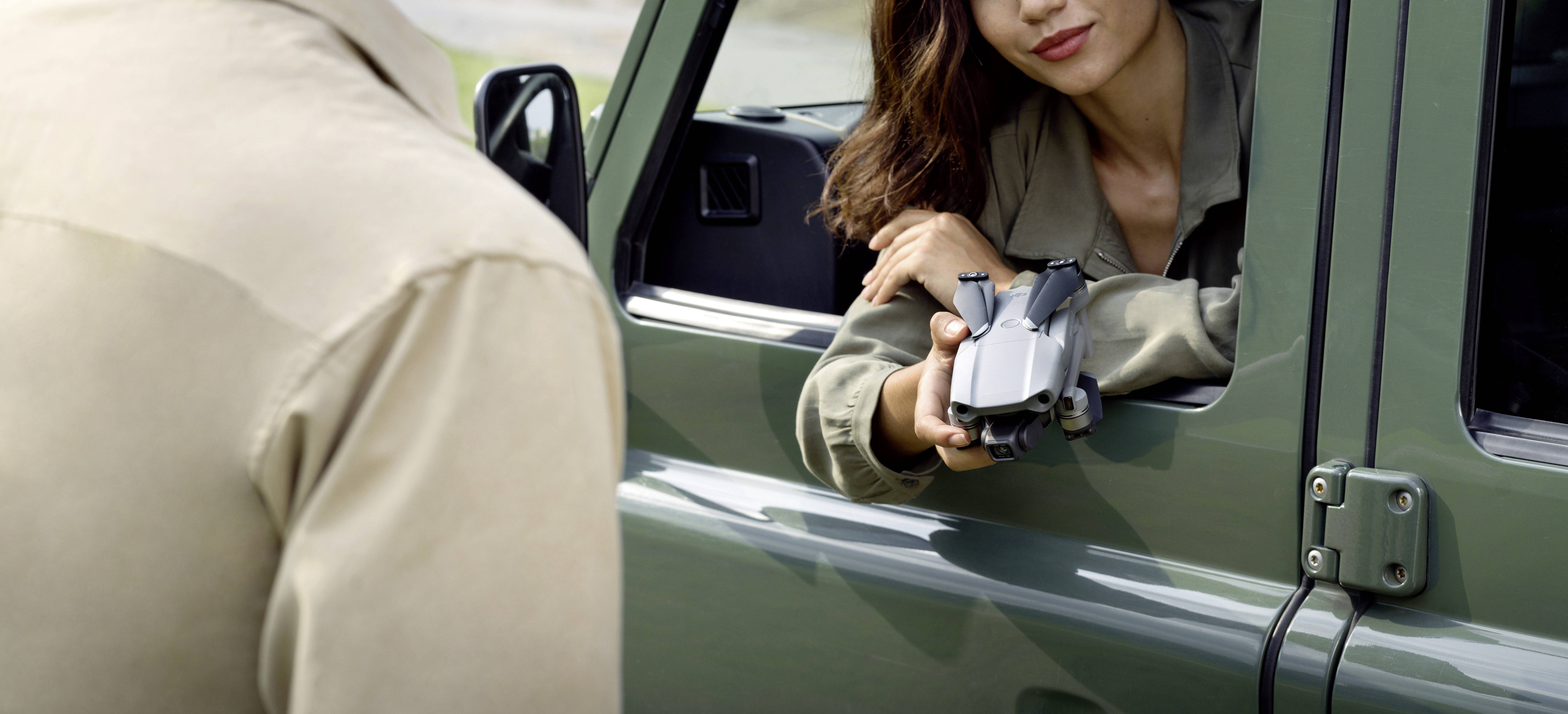 Eine Frau in einem Jeep reicht einem stehenden Mann eine zusammengefaltete Drohne durchs Fenster. Beide wirken konzentriert.