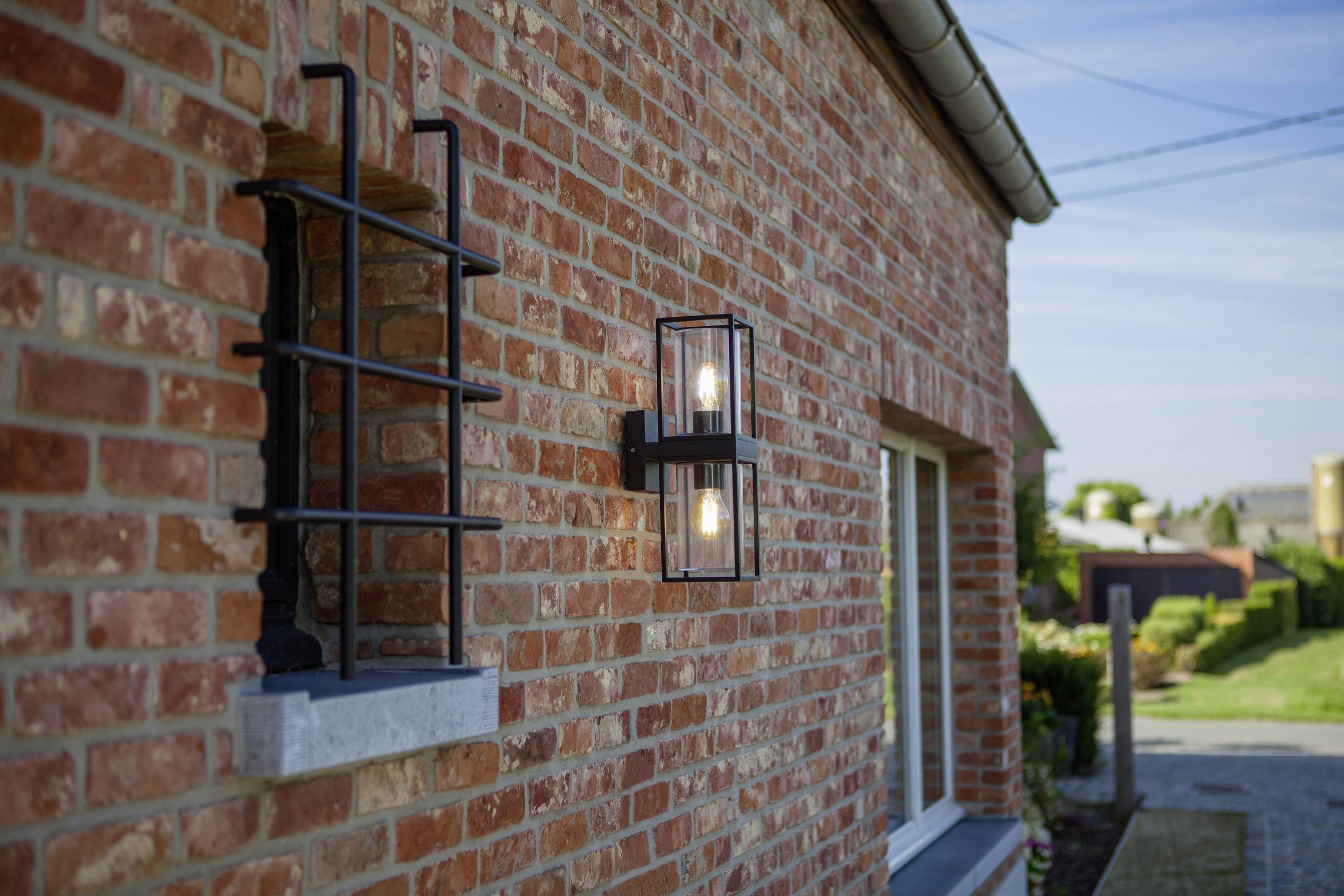 Eine rote Backsteinmauer mit einem angebrachten Wandlicht aus Metall und Glas. Im Hintergrund ist ein grüner Garten zu sehen.