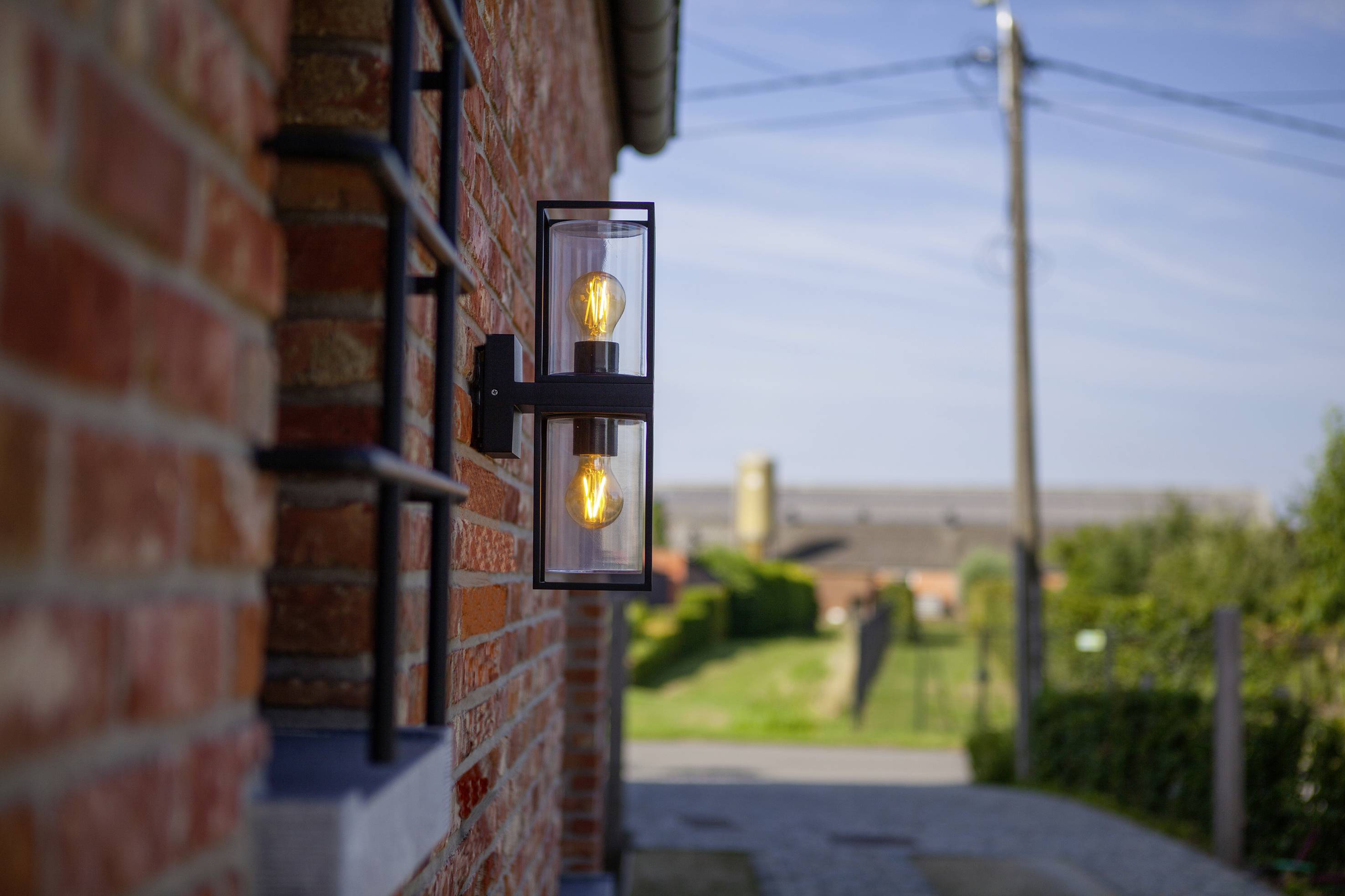 Eine moderne Außenwandlampe leuchtet an einer Backsteinmauer. Im Hintergrund ist eine ländliche Straße mit Strommasten zu sehen.