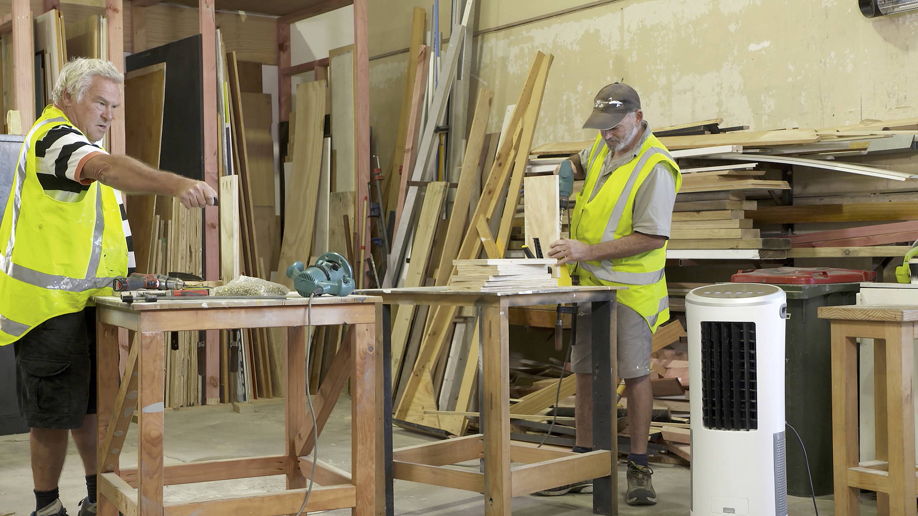 Zwei Arbeiter in gelben Warnwesten arbeiten in einer Holzwerkstatt, umgeben von Holzstapeln und Werkzeugen auf einem Tisch.