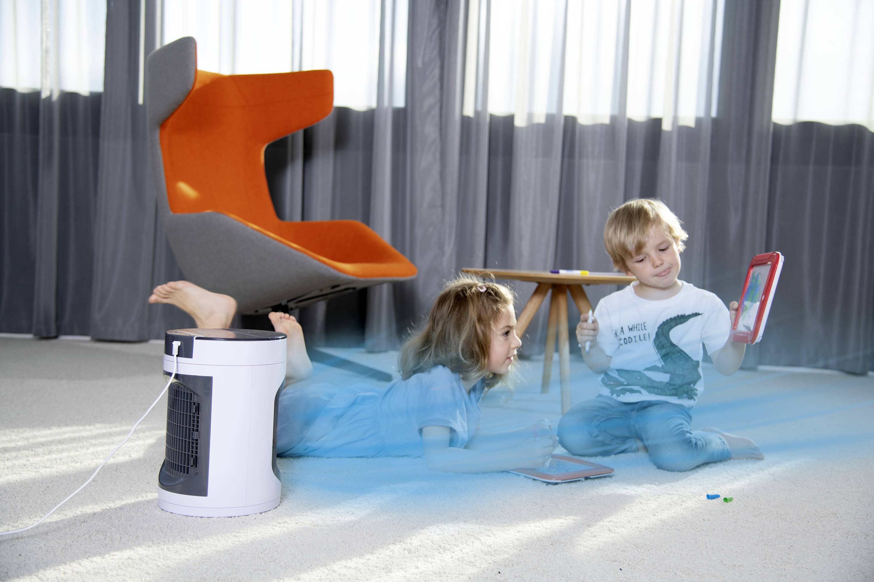 Zwei Kinder im Wohnzimmer. Ein Kind liegt auf dem Bauch und schaut auf ein Tablet. Ein Luftreiniger steht auf dem Boden. Ein moderner Stuhl im Hintergrund.