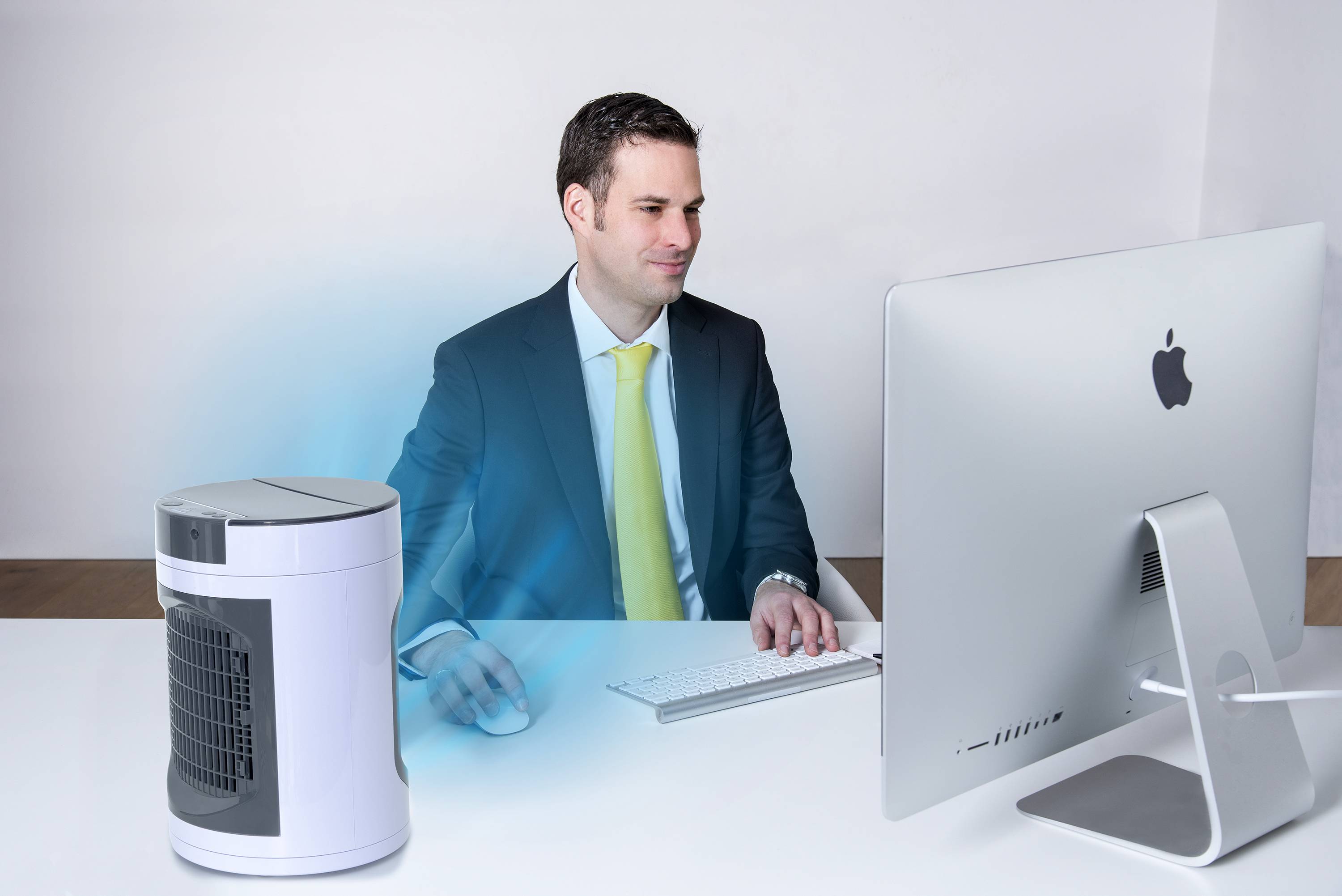 Ein Mann in Anzug sitzt an einem Schreibtisch vor einem Computer und arbeitet konzentriert, während ein Luftreiniger in der Nähe steht.