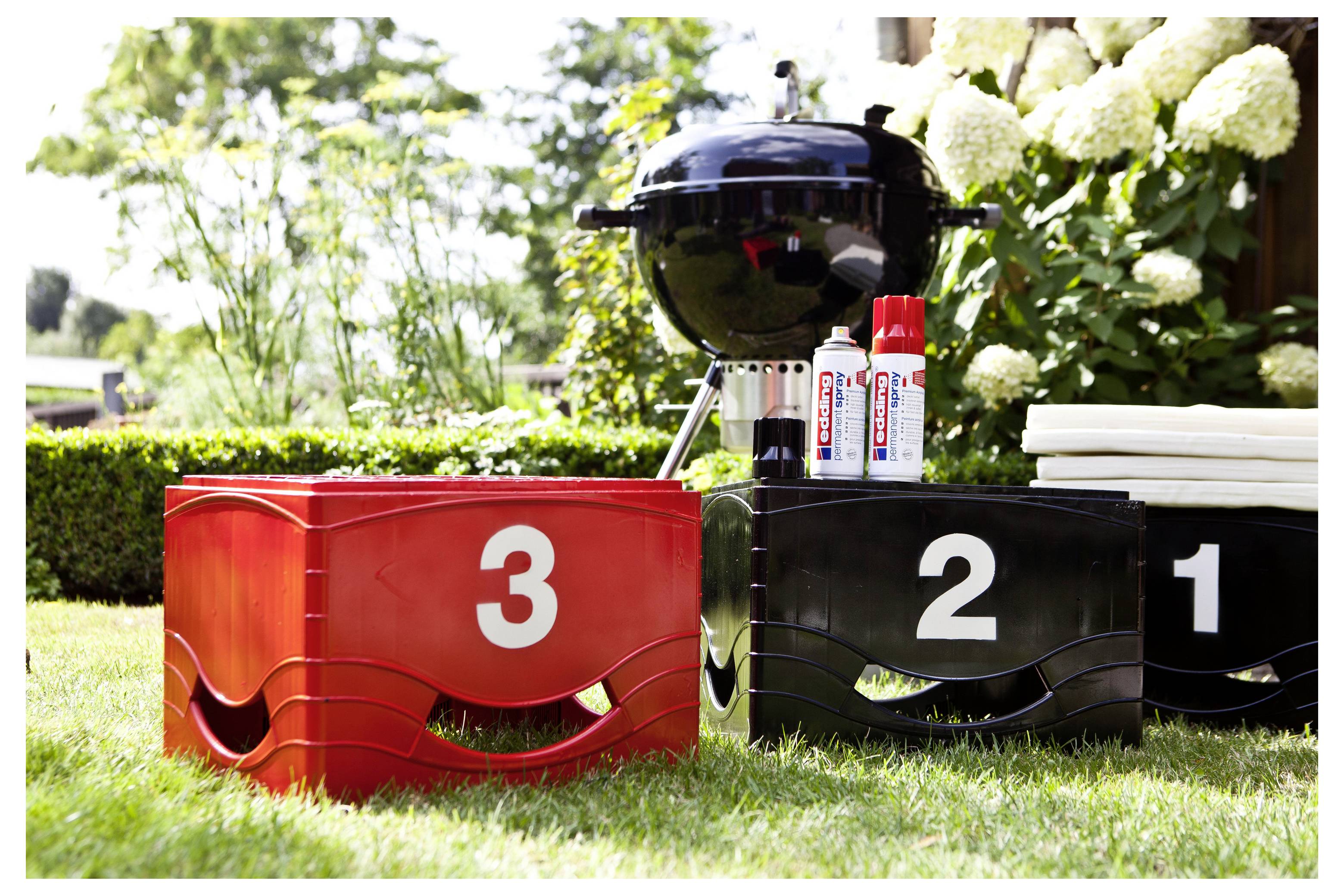 Outdoor-Grillaufbau mit einem schwarzen Grill und gestapelten schwarzen, roten und weißen nummerierten Behältern mit den Nummern 3, 2 und 1 auf Gras.