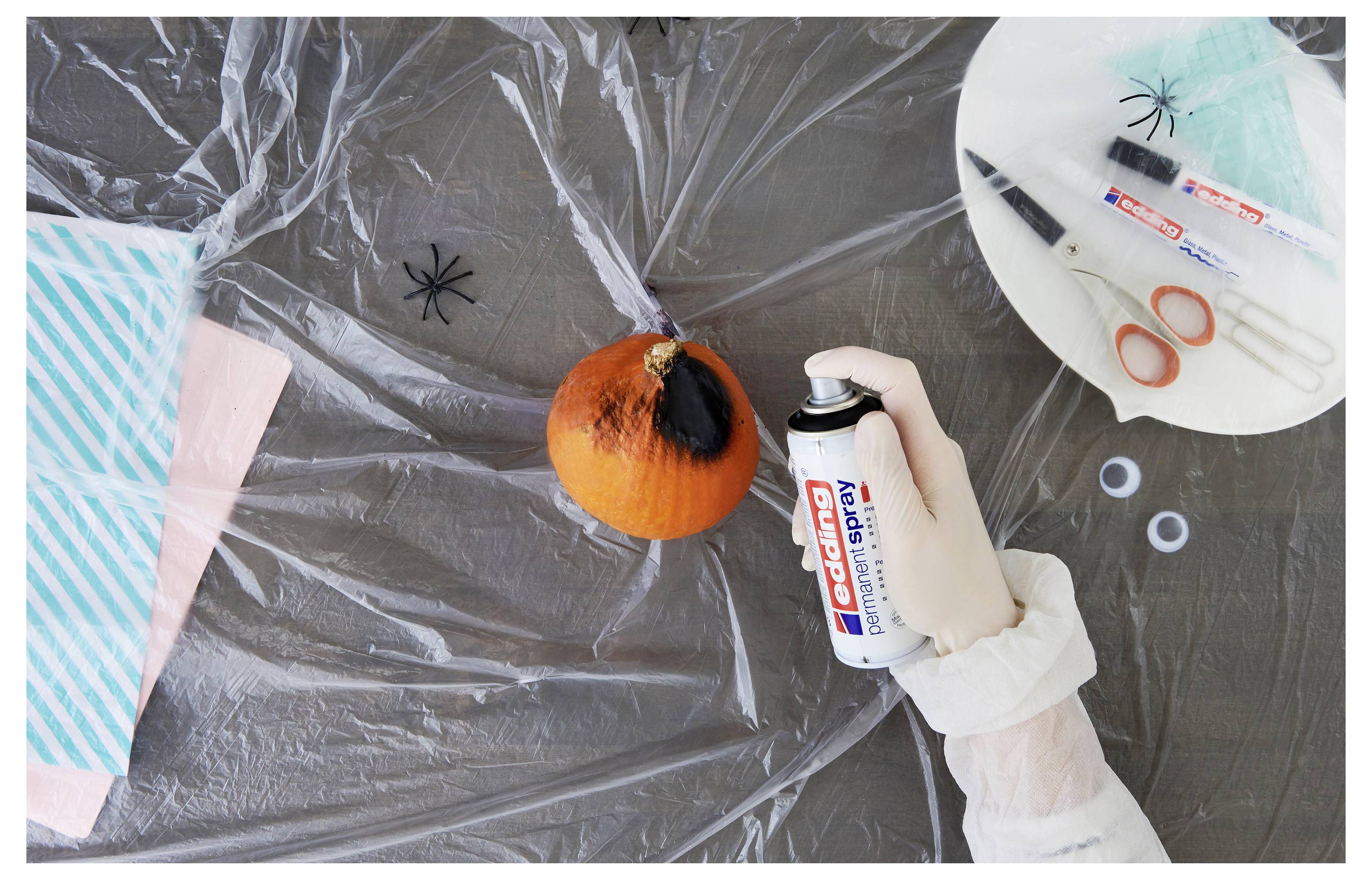 Hand in Handschuh sprüht weiße Farbe auf einen kleinen orangefarbenen Kürbis auf einem mit Kunststoff abgedeckten Tisch, mit Markern, Schere und Papier in der Nähe.