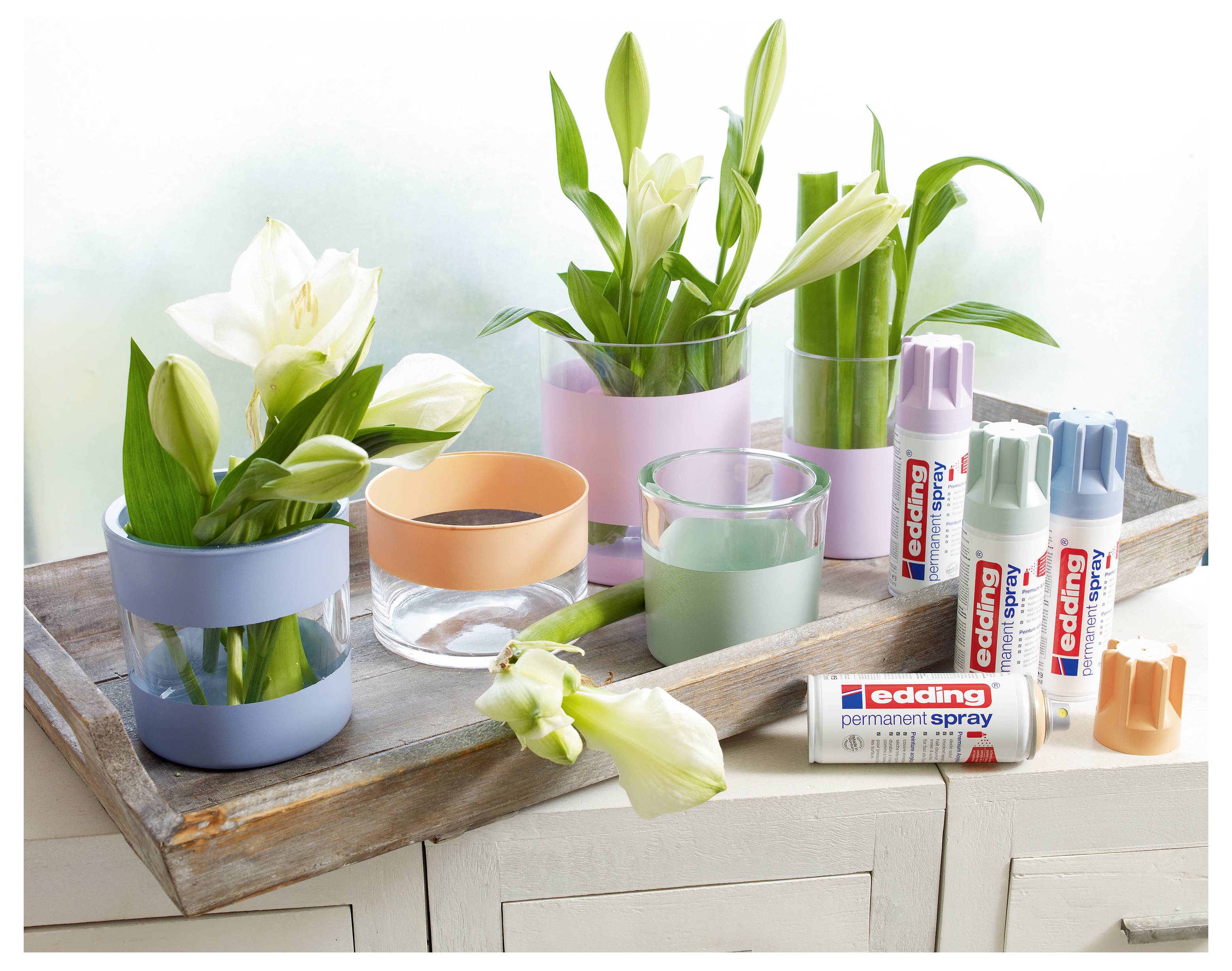 Dekorative Blumenarrangement in Pastellgläsern auf einem Holztablett, mit mehreren "edding"-Permanent-Sprühdosen und Farbkappen daneben