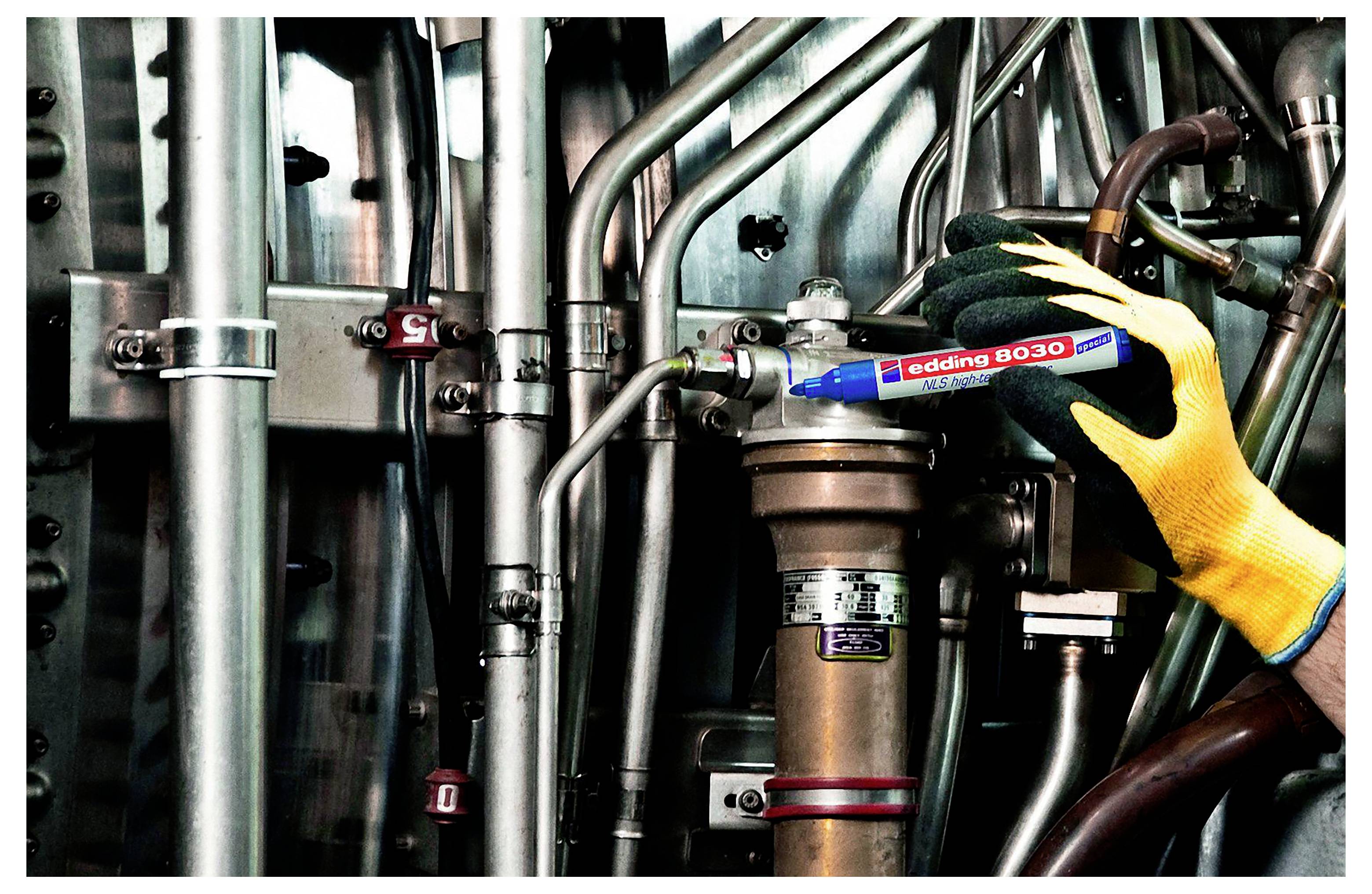 Behandschuhte Hand beschriftet Industrierohre mit einem blauen „edding 830"-Marker in einem Maschinenraum.