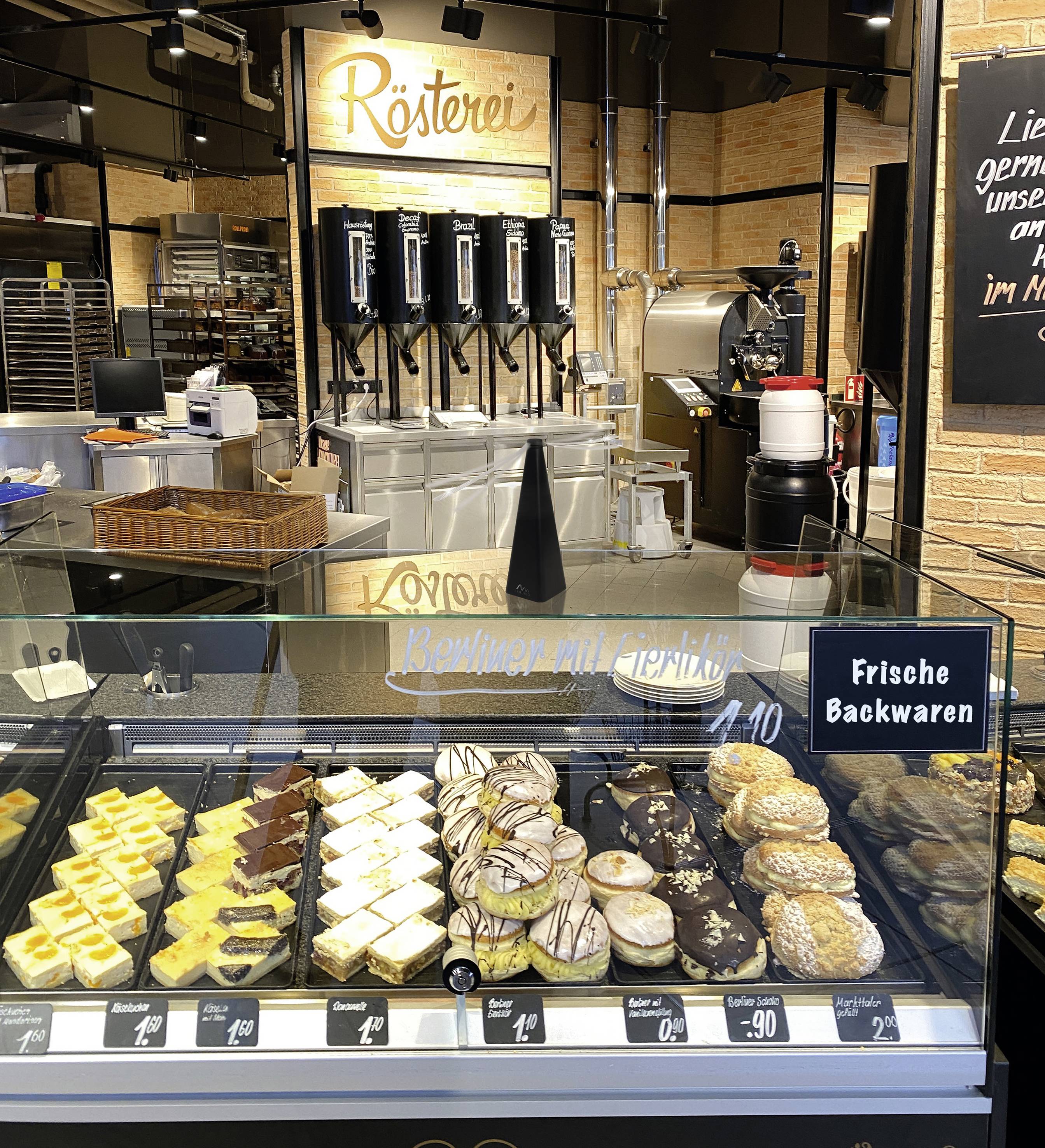 Eine Bäckertheke mit Auswahl an Gebäck, darunter Kuchenstücke und Donuts. Im Hintergrund steht 'Rösterei' über mehreren Kaffeeautomaten.