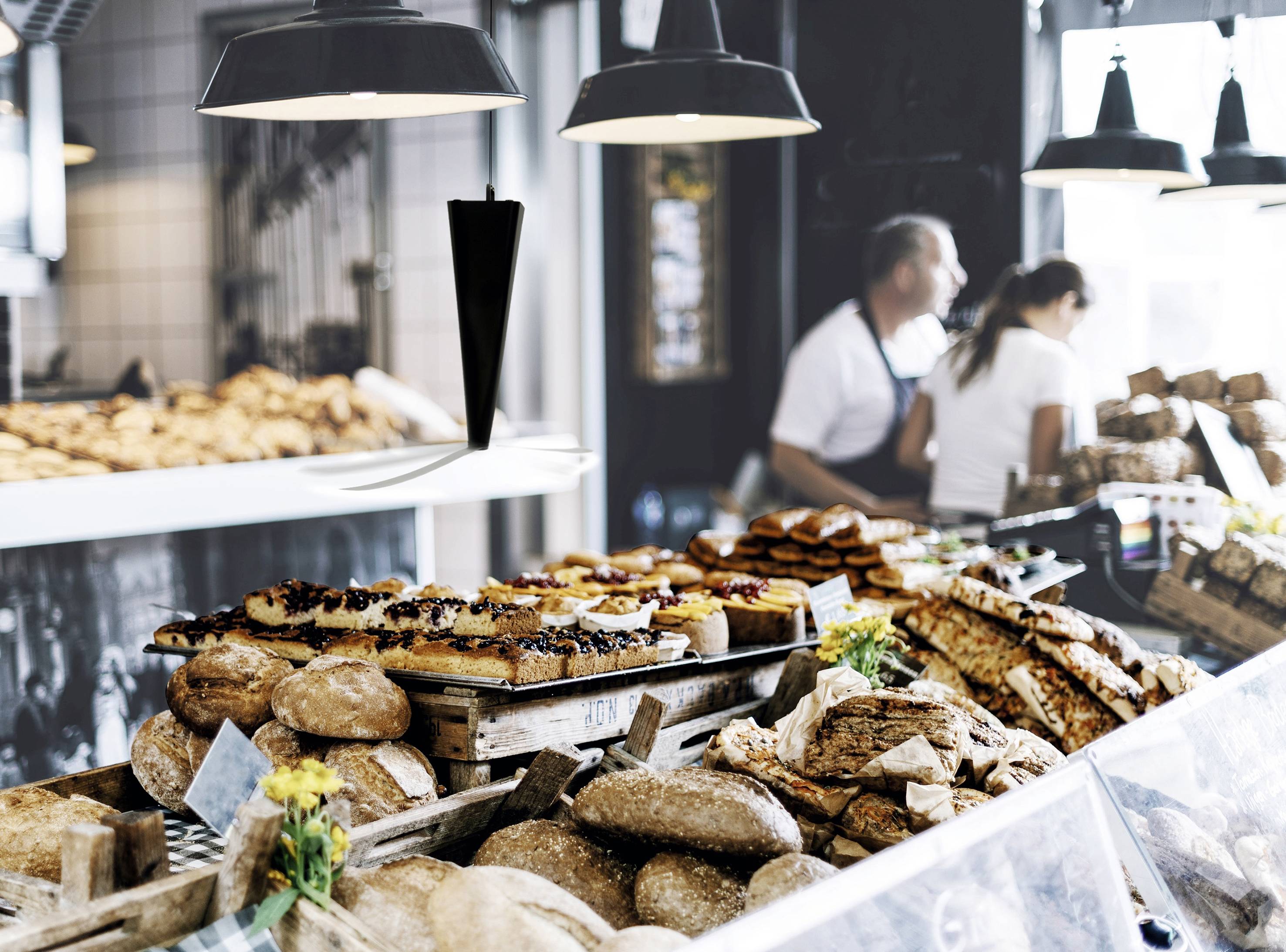 Eine Bäckerei mit einer Auswahl an Broten und Gebäck auf Theken. Im Hintergrund arbeiten zwei Personen.sprechende Bekleidung.