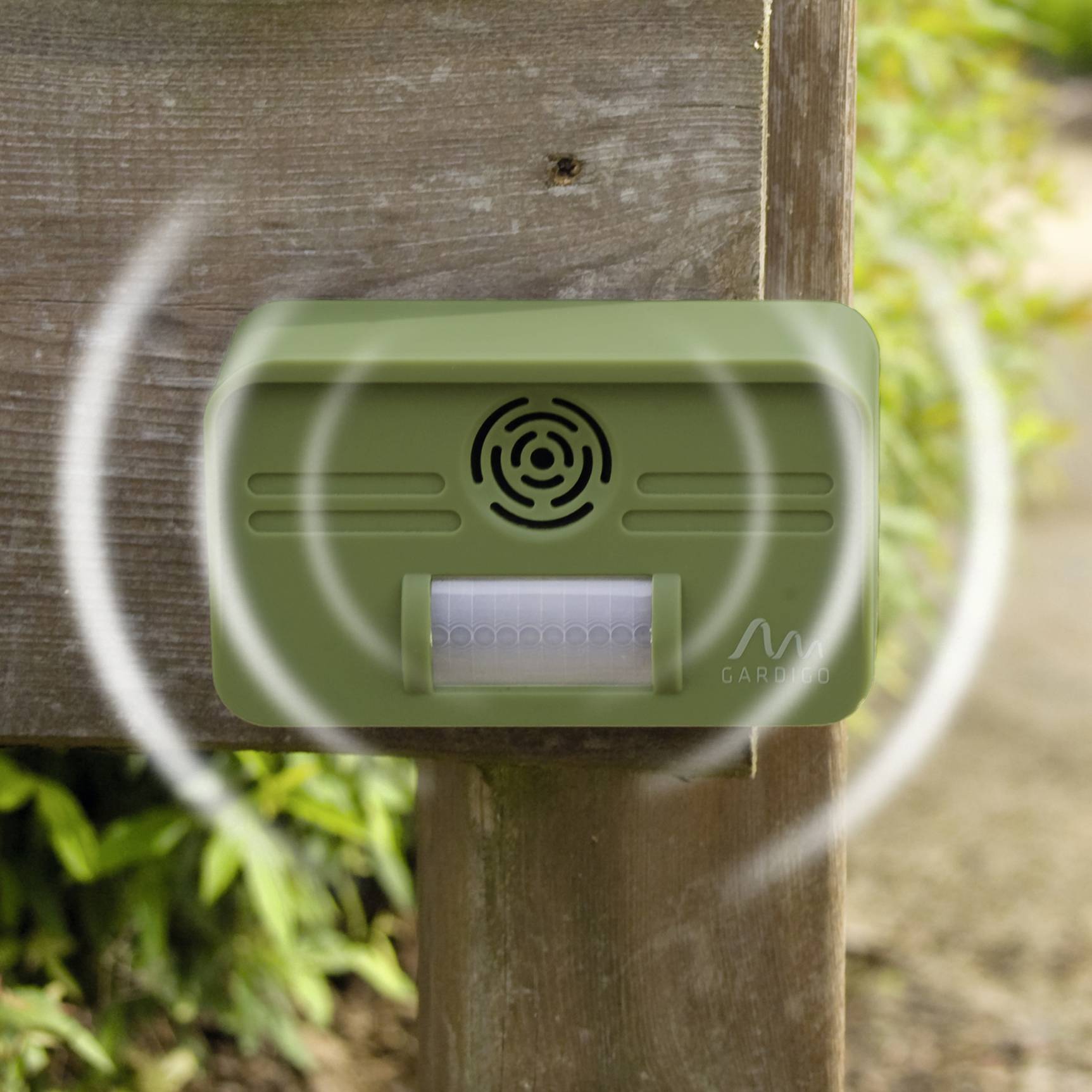 Ein grünes Ultraschall-Tiervertreibergerät, montiert an einem Holzpfosten im Garten, sendet Schallwellen zur Abschreckung aus.