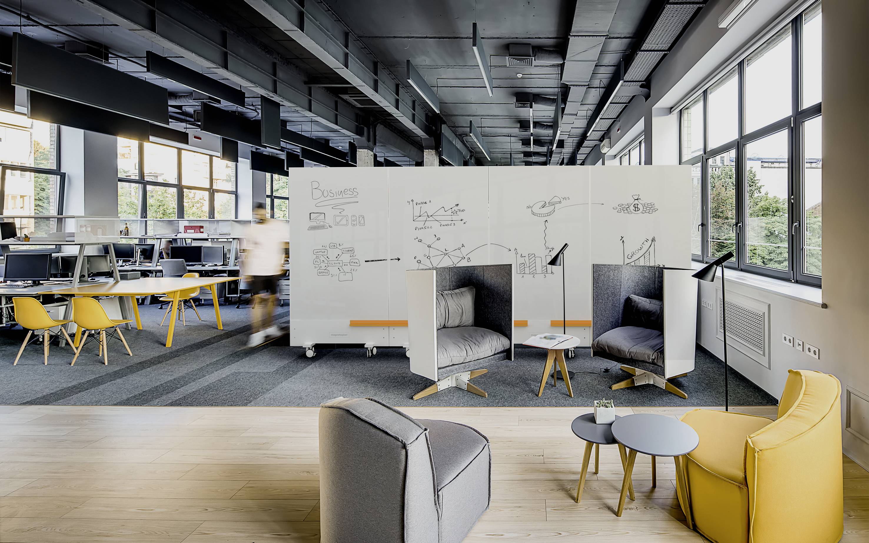 Ein modernes Büro mit offenen Arbeitsbereichen, Sitzgelegenheiten, Whiteboards und großen Fenstern. Personen arbeiten und bewegen sich im Raum.