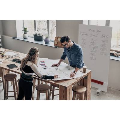 Zwei Personen arbeiten an einem großen Papier auf einem Holztisch in einem modernen Büro. Im Hintergrund ist ein Projektplan an einem Whiteboard sichtbar.
