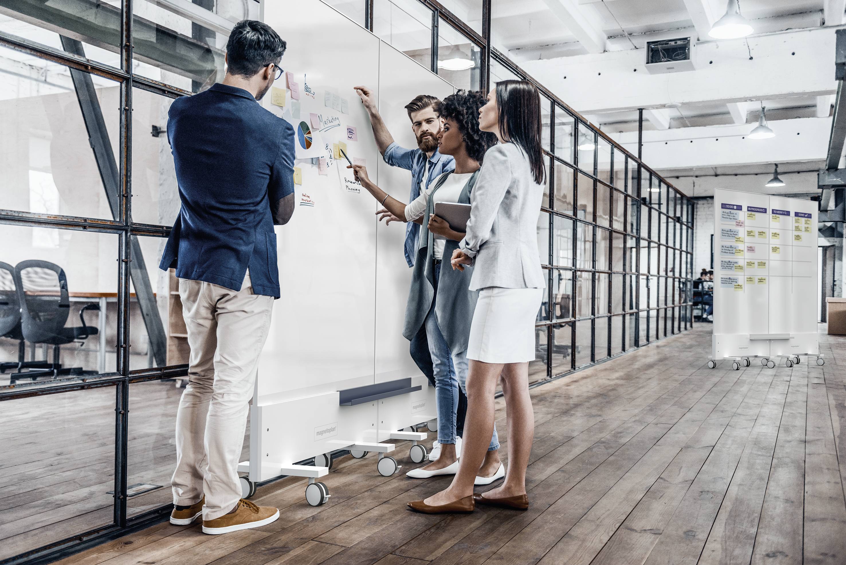 Vier Personen befinden sich in einem modernen Büro und diskutieren an einem Whiteboard. Sie zeigen auf Notizen und Diagramme.
