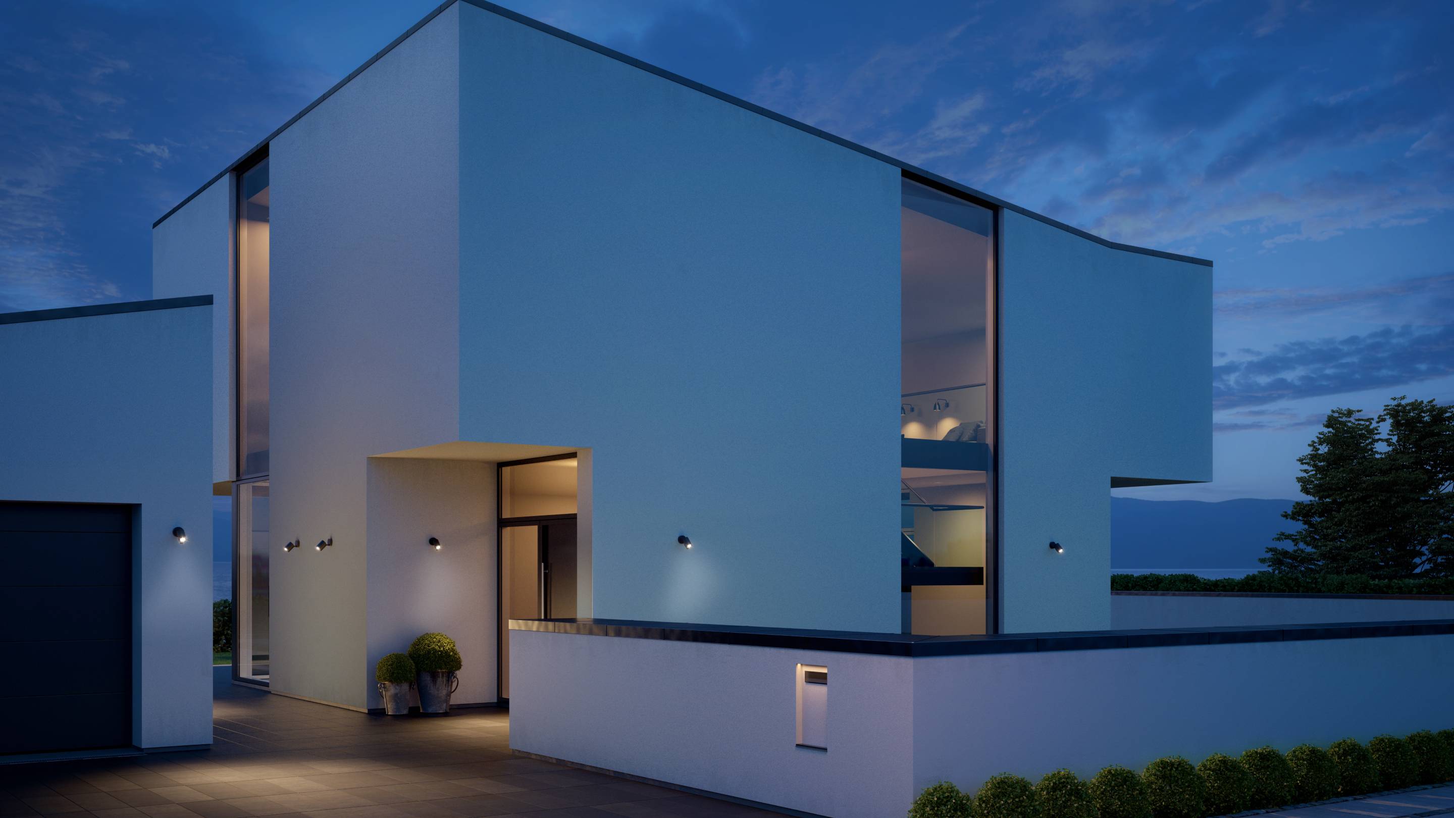 Modernes zweistöckiges Haus mit Flachdach, großflächigen Fenstern und umgeben von einem Garten. Die Beleuchtung im Haus ist an.