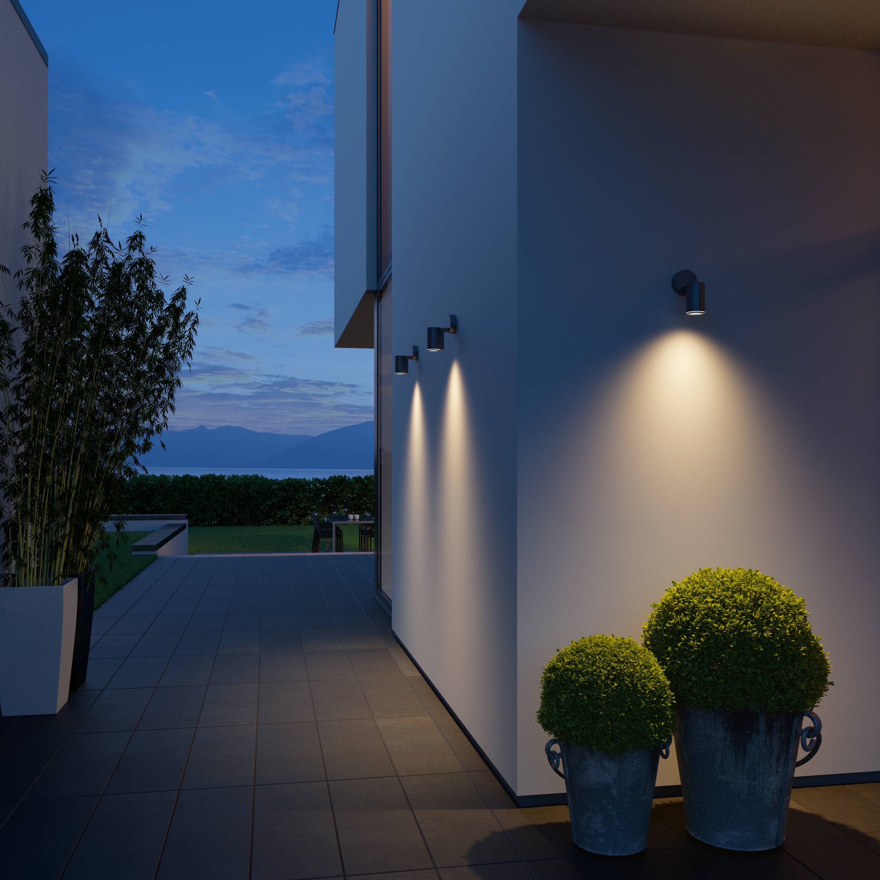 Moderne Architektur bei Abenddämmerung mit beleuchteter Außenwand. Im Vordergrund zwei grüne Büsche in Töpfen, dahinter Meerblick.