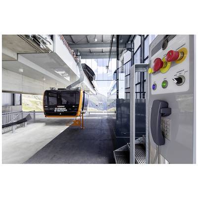 Eine Gondelkabine in einer modernen Bergstation, umgeben von großen Fenstern, bietet einen Blick auf die Berge im Hintergrund. Ein Bedienfeld ist im Vordergrund zu sehen.