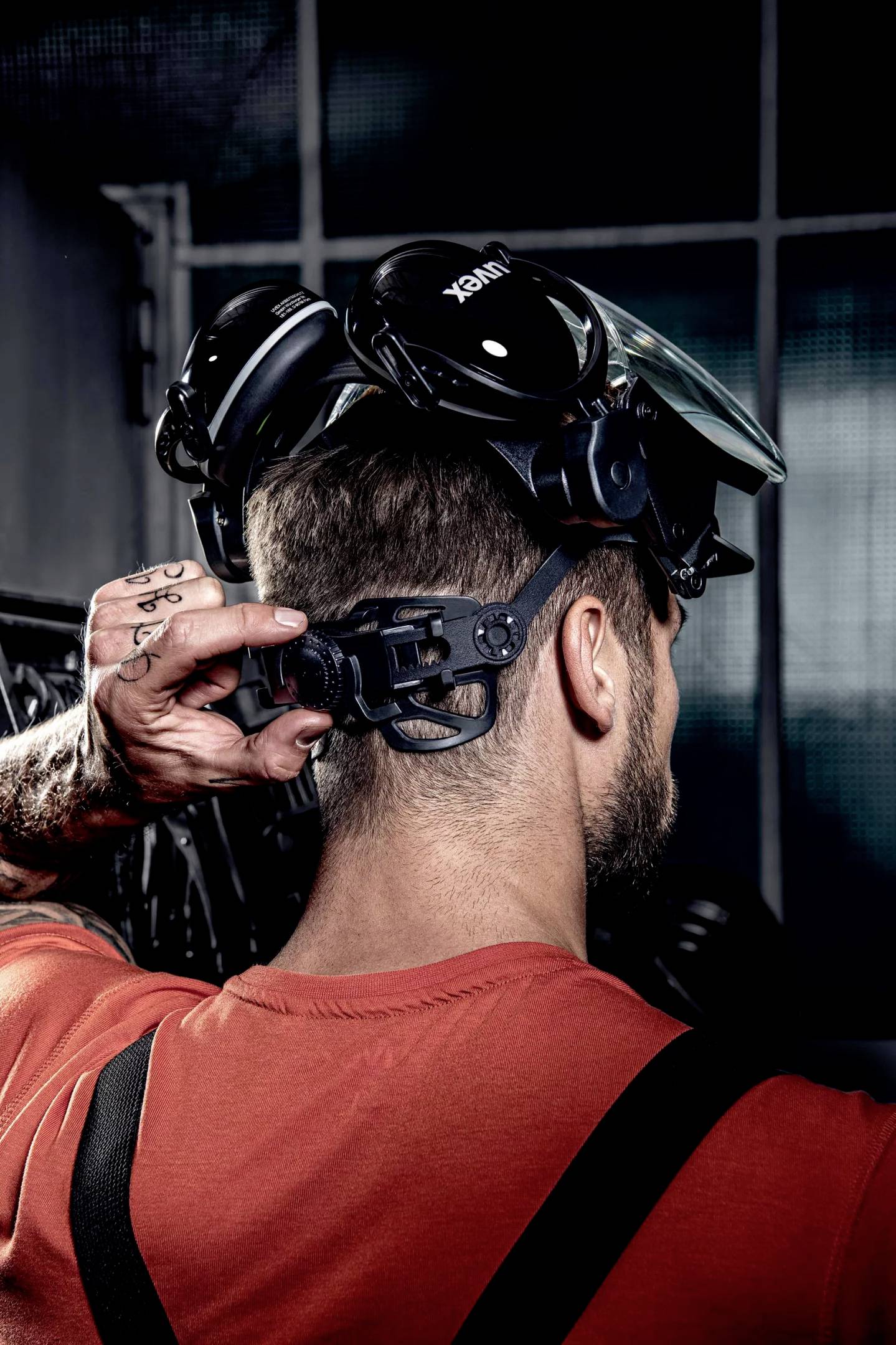 Ein Mann in einem roten T-Shirt trägt eine Schutzbrille und Gehörschutz auf dem Kopf. Er steht in einer industriellen Umgebung.