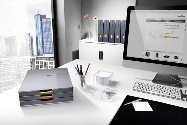 Ein modernes Büro mit einem Schreibtisch, auf dem ein Computer, eine Tastatur, Schreibutensilien und ein Ordnersystem stehen. Im Hintergrund ist eine Stadtansicht zu sehen.