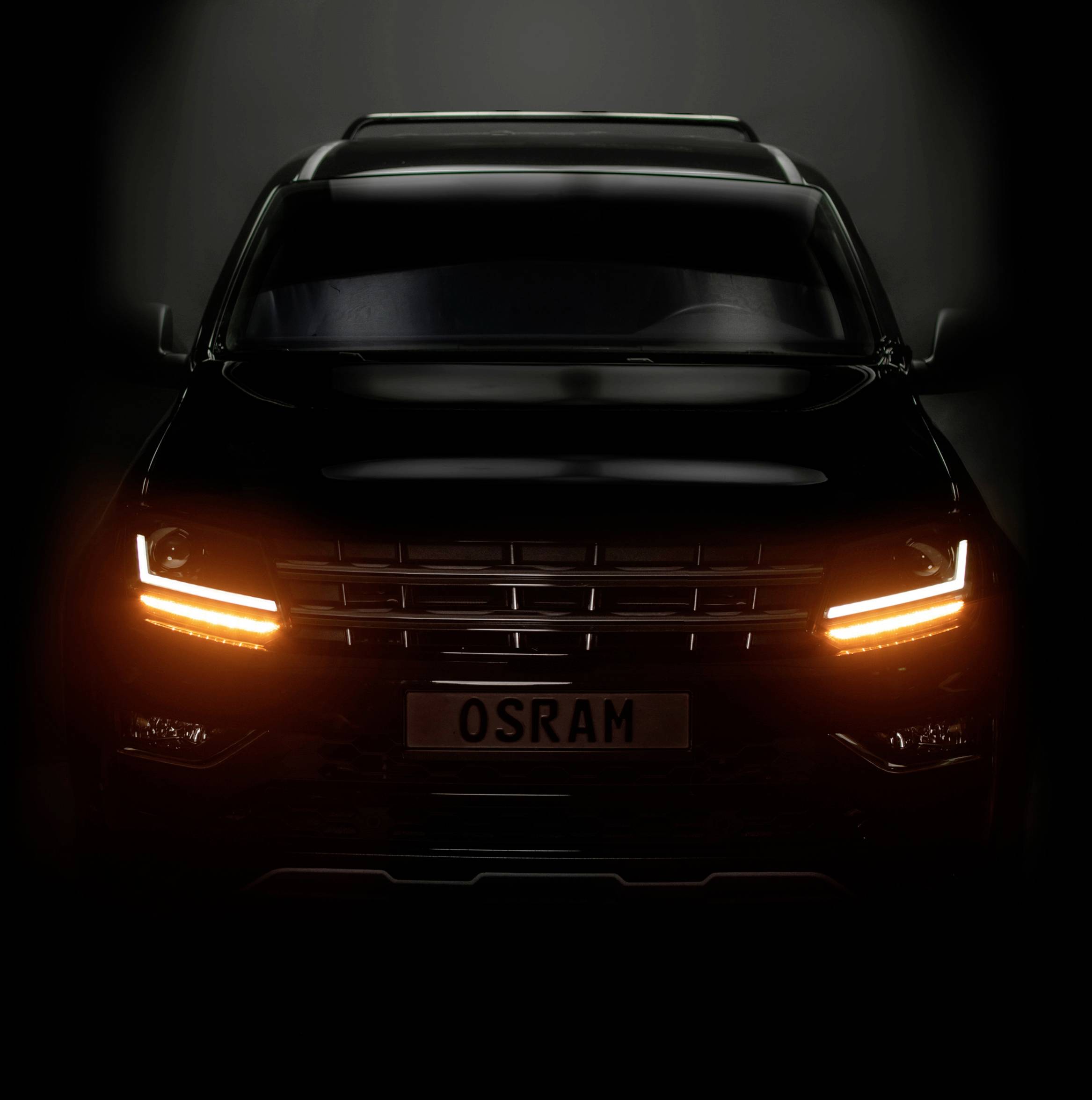 Ein schwarzes Auto mit leuchtenden Frontscheinwerfern, das frontal im Dunkeln gezeigt wird. Auf dem Nummernschild steht 'OSRAM'.