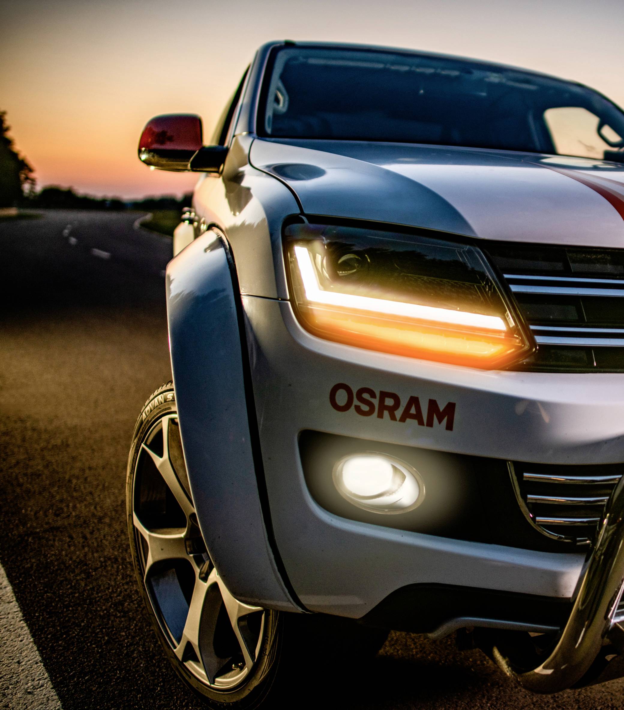 Ein weißer SUV mit eingeschaltetem Frontlicht und einem OSRAM-Logo steht bei Sonnenuntergang auf einer Landstraße.