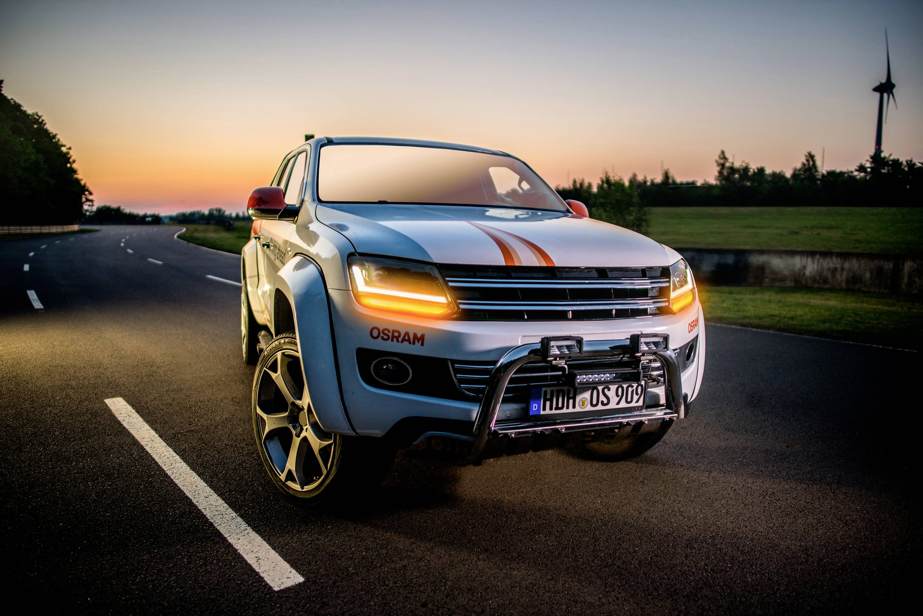 Ein weißes Auto mit goldenen Felgen steht auf einer Landstraße bei Sonnenuntergang. Im Hintergrund ist ein Windrad sichtbar.