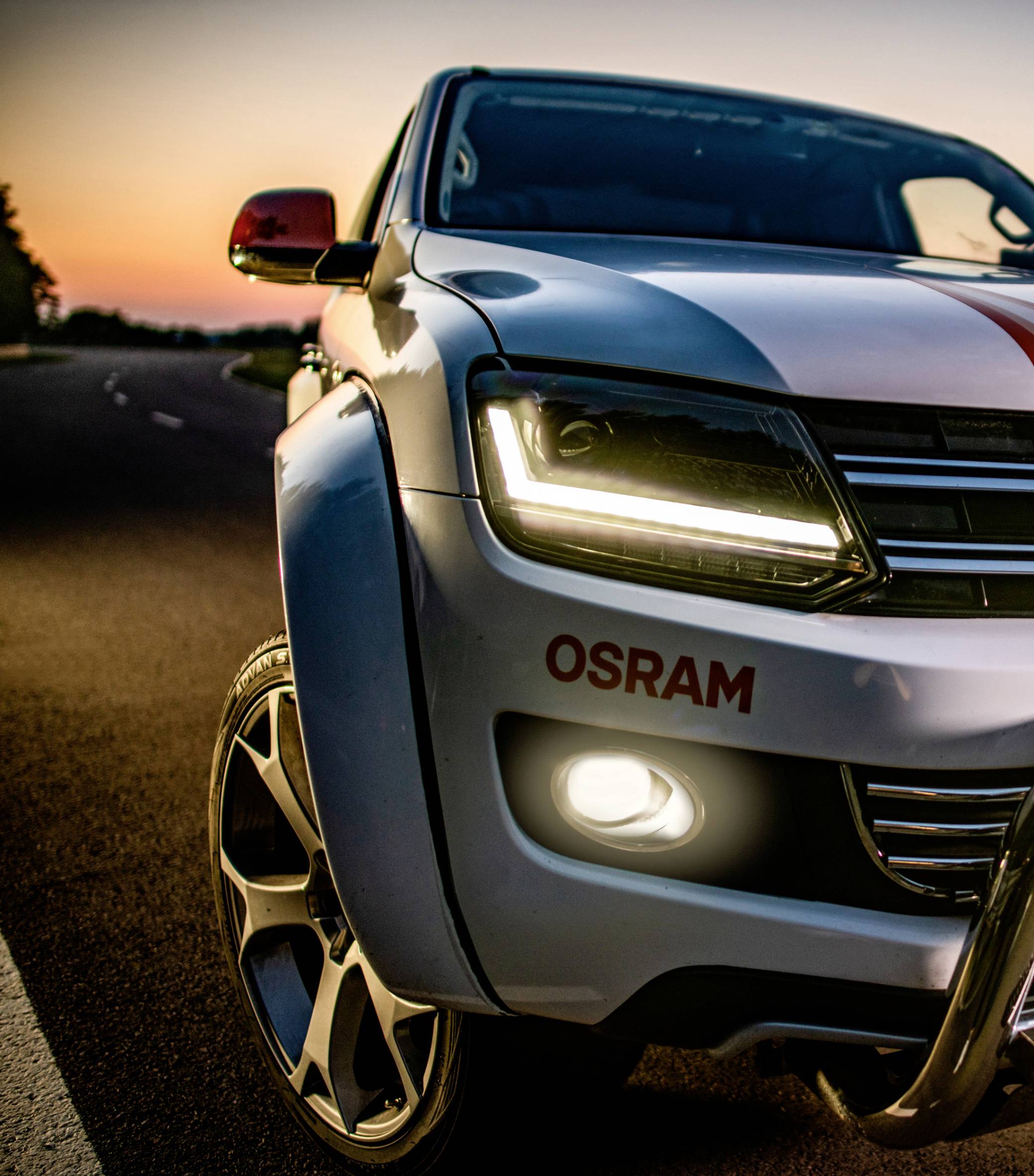 Ein Auto fährt bei Sonnenuntergang auf einer Straße. Der Scheinwerfer ist eingeschaltet. Auf dem Auto steht 'OSRAM'.