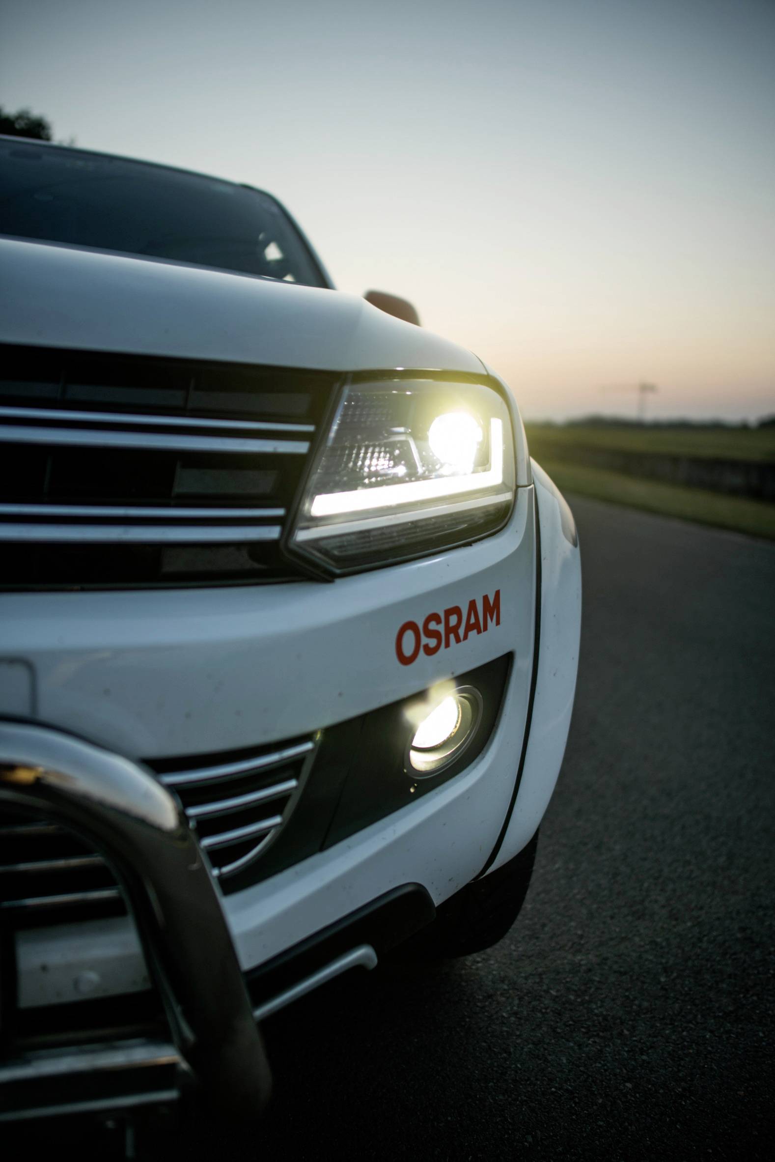 SUV mit eingeschaltetem Scheinwerfer und 'OSRAM'-Aufschrift, bei Sonnenuntergang auf einer Straße geparkt.