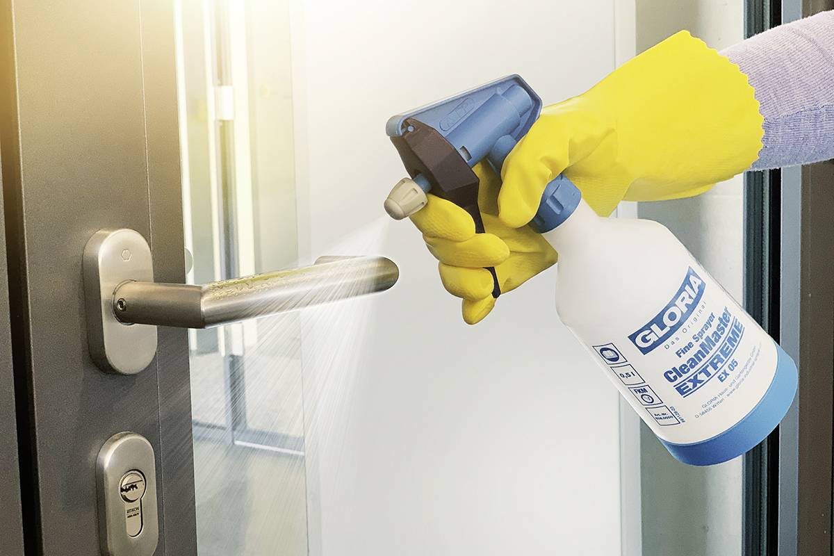 Eine Hand mit gelbem Handschuh hält einen Sprühreiniger und desinfiziert einen Türgriff in einem modernen Gebäude.