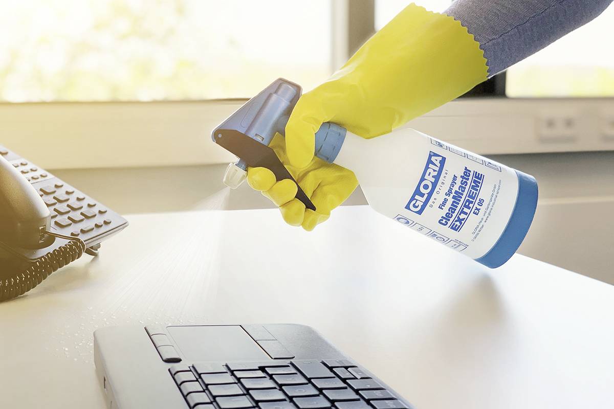 Eine Person reinigt eine Computer-Tastatur mit einem Desinfektionsspray, während sie gelbe Gummihandschuhe trägt.