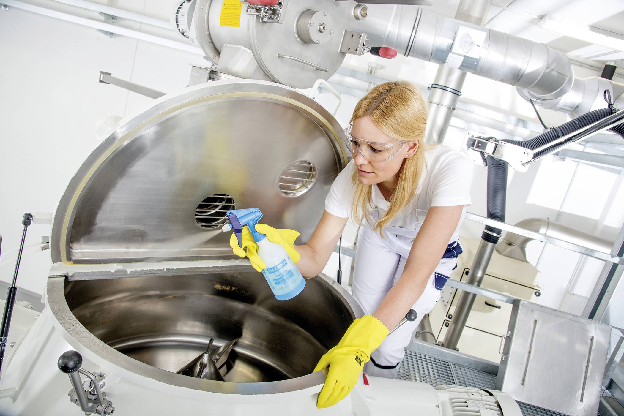 Eine Frau in Schutzkleidung reinigt eine große Metallmaschine mit einem blauen Sprühmittel in einer industriellen Umgebung.