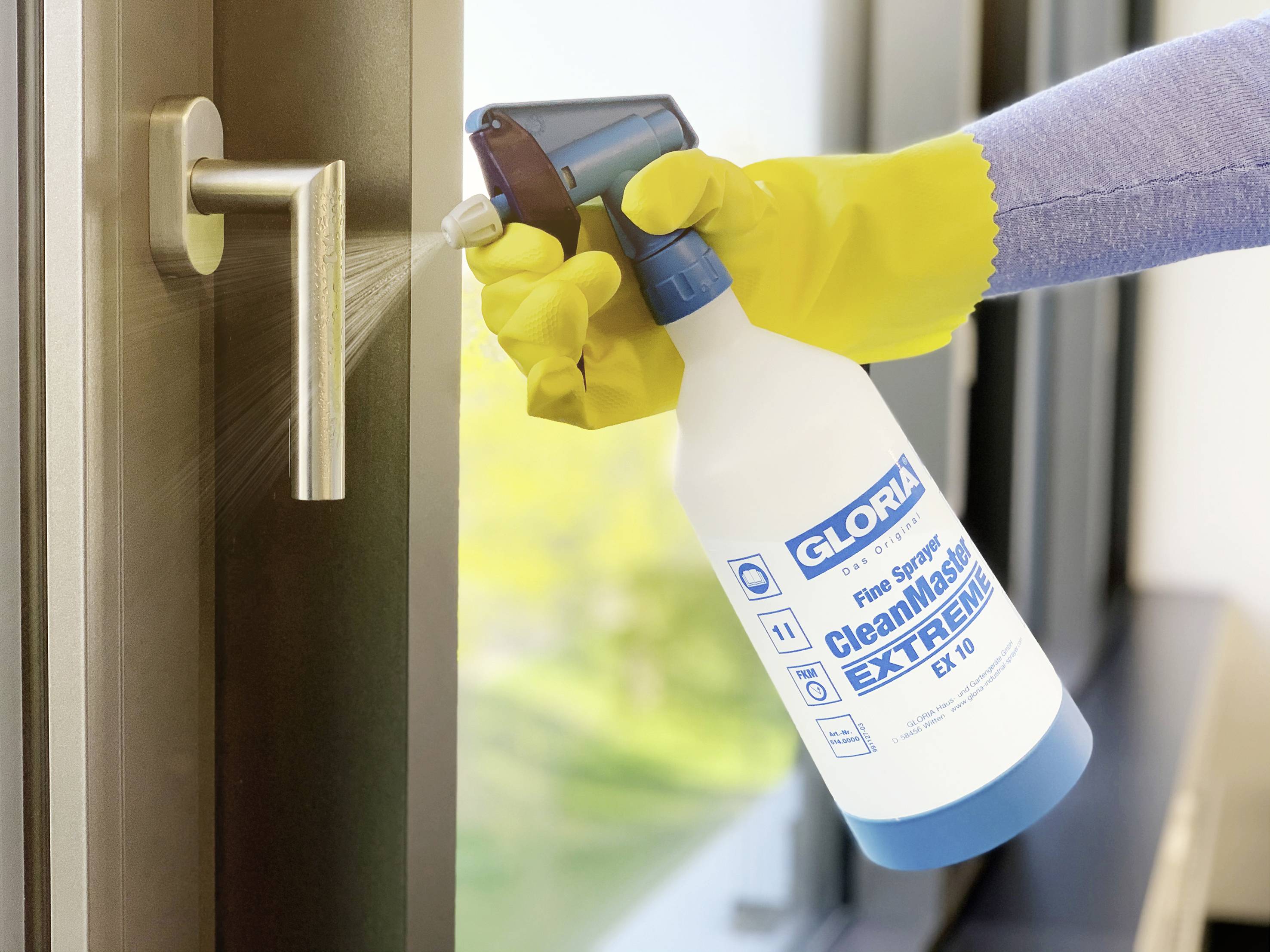 Hand mit gelbem Handschuh sprüht Reinigungsmittel aus einer Flasche auf einen Türgriff, um ihn zu desinfizieren.