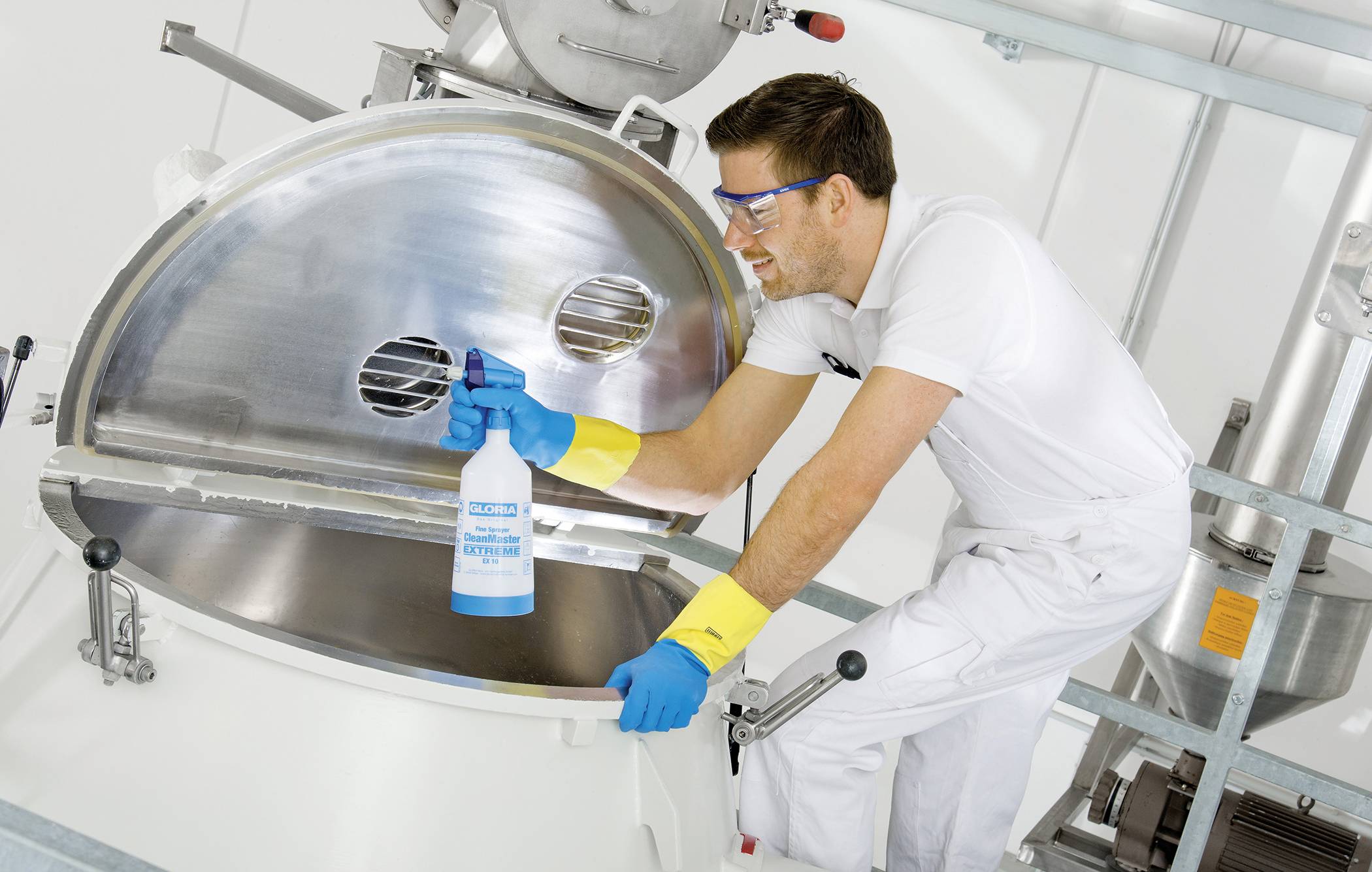 Ein Mann reinigt mit Schutzbrille, Handschuhen und Reinigungsmittel eine große Metallmaschine in einer industriellen Umgebung.
