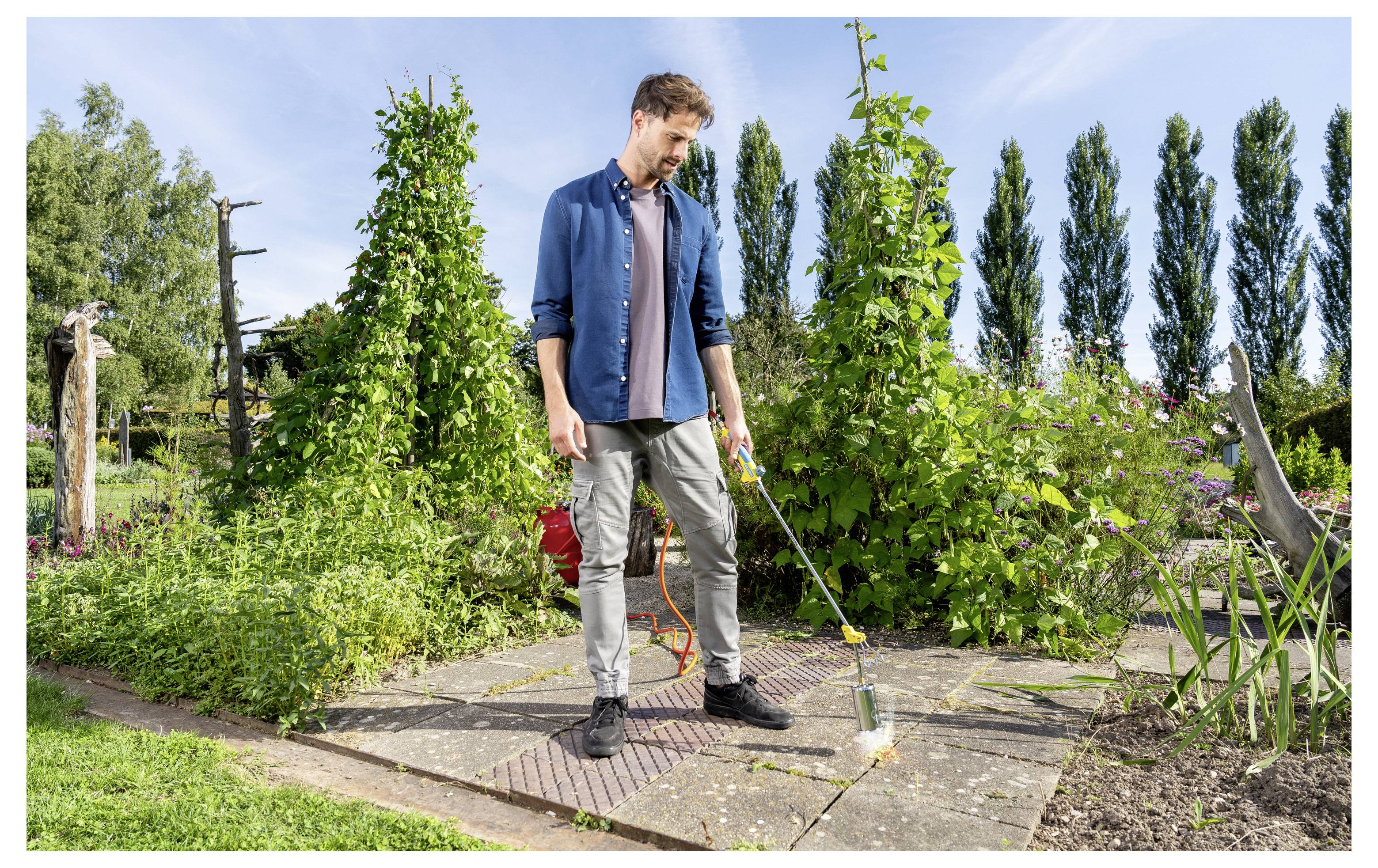 Ein Mann benutzt einen Unkrautbrenner auf einem gepflasterten Gartenweg neben üppig grünen Pflanzen und Sträuchern an einem sonnigen Tag.