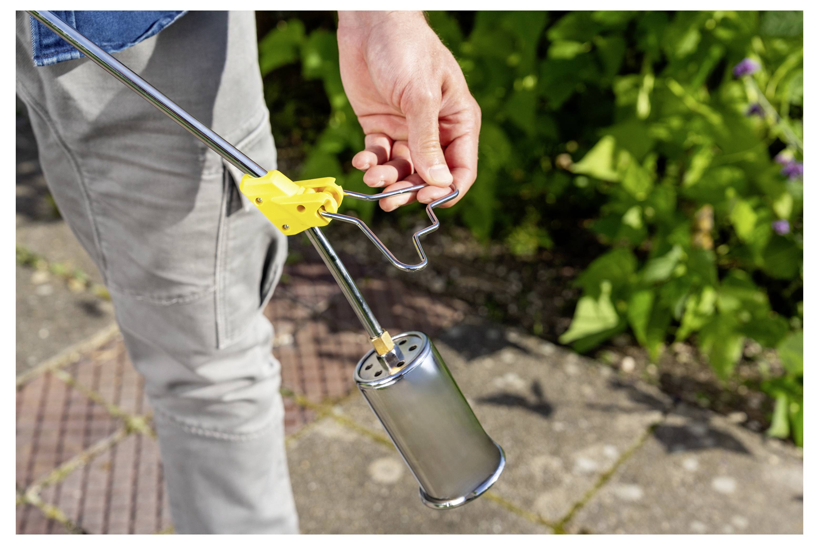 Person, die einen Metalunkrautvernichter-Fackel mit gelbem Zündaufsatz im Freien hält, auf den Boden neben einem Gartenweg gerichtet.