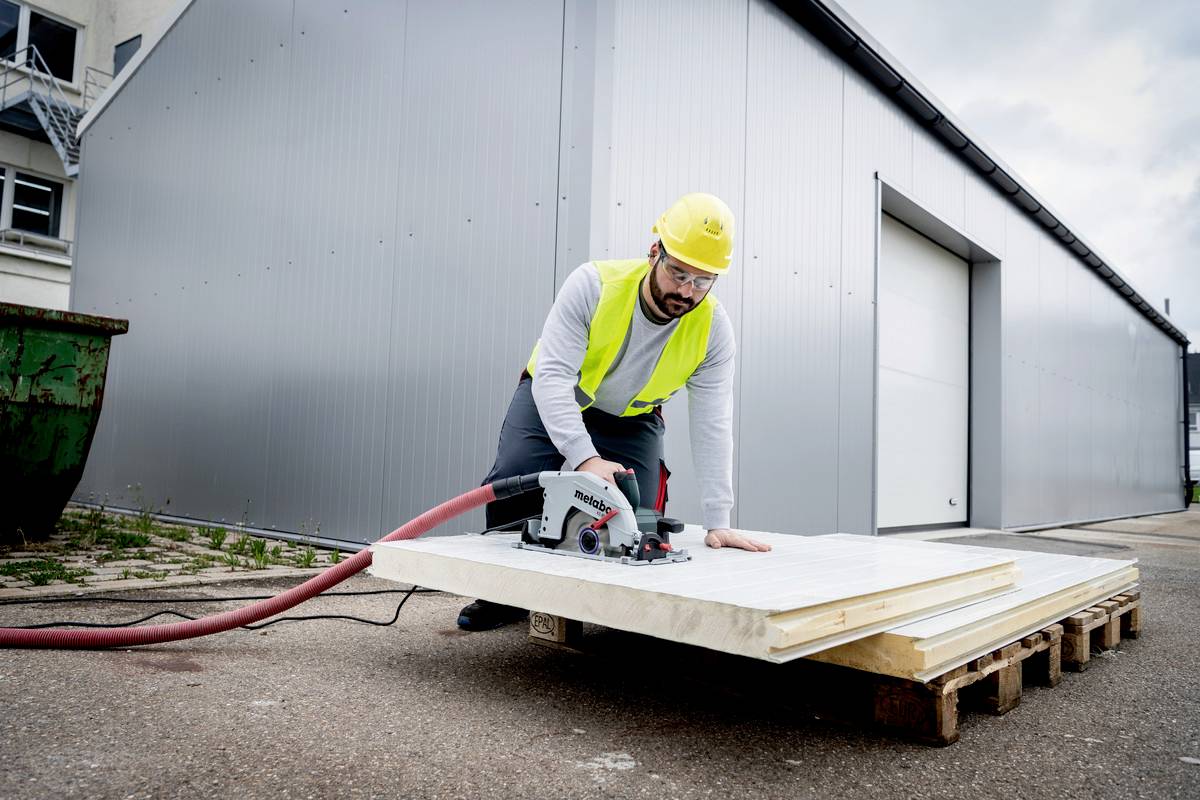 Arbeiter mit Schutzhelm und Weste schneidet mit Kreissäge Holzplatte auf Palette vor Lagerhalle.