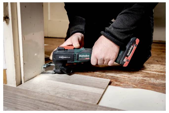 Eine Person benutzt ein Metabo-Elektrowerkzeug, um ein Stück Holzboden in der Nähe eines Türrahmens zu schneiden.