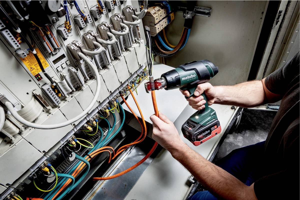 Eine Person arbeitet mit einem Akkuschrauber an einem elektrischen Schaltschrank. Mehrere Kabel und Komponenten sind sichtbar.