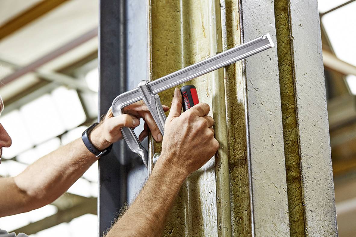 Eine Person misst mit einer großen silbernen Klammermaßstange einen Metallbalken in einem industriellen Umfeld.