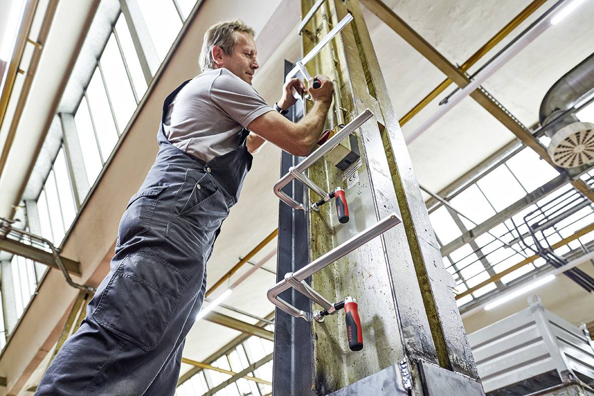 Ein Handwerker arbeitet mit Werkzeugen an einem Stahlträger in einer großen, hellen Werkstattumgebung.